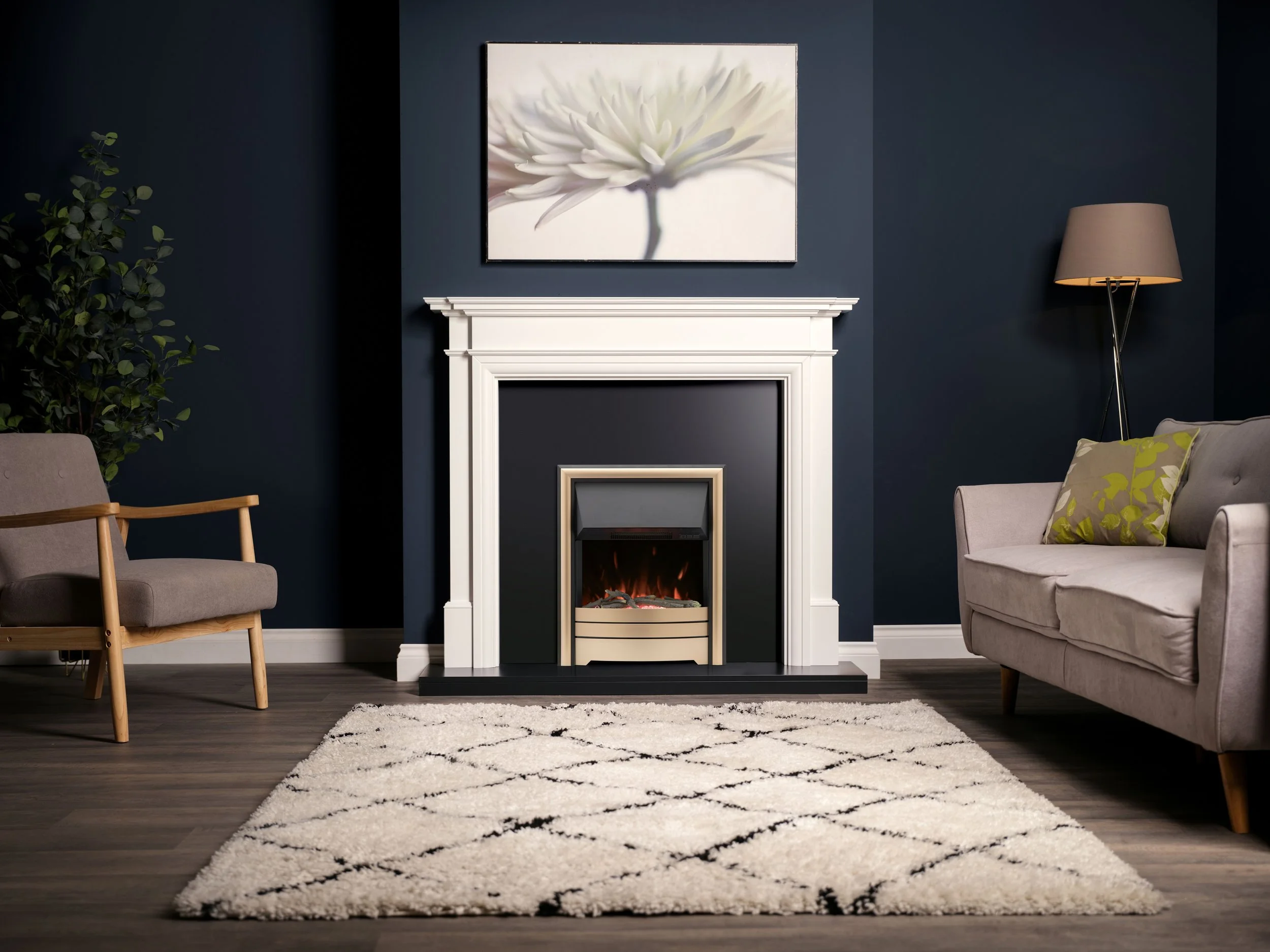 Living room with white fireplace, navy walls, grey sofa with green patterned pillow, grey armchair, beige lamp, abstract floral artwork, potted plant, white area rug, dark wood floor.