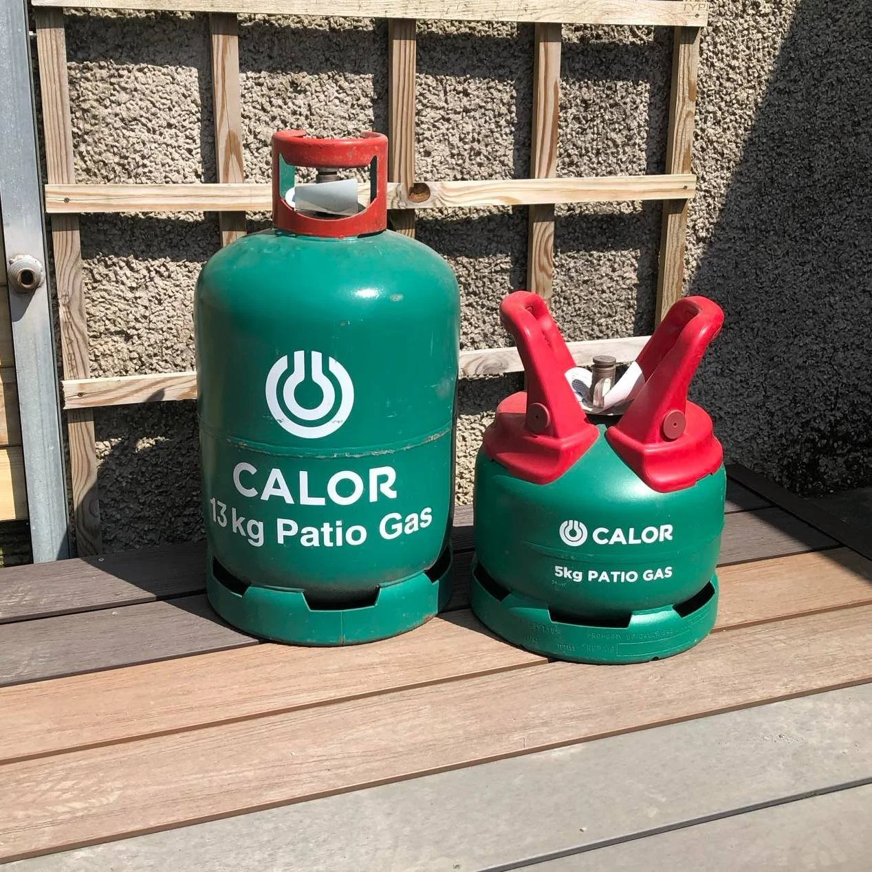 Two green patio gas cylinders labeled 'Calor,' one 13 kg and the other 5 kg, are placed on a wooden outdoor surface against a background of a fenced wall and a wall with textured stucco.