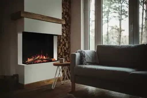 Cozy living room with fireplace, wooden logs, gray sofa, and large window with trees outside.