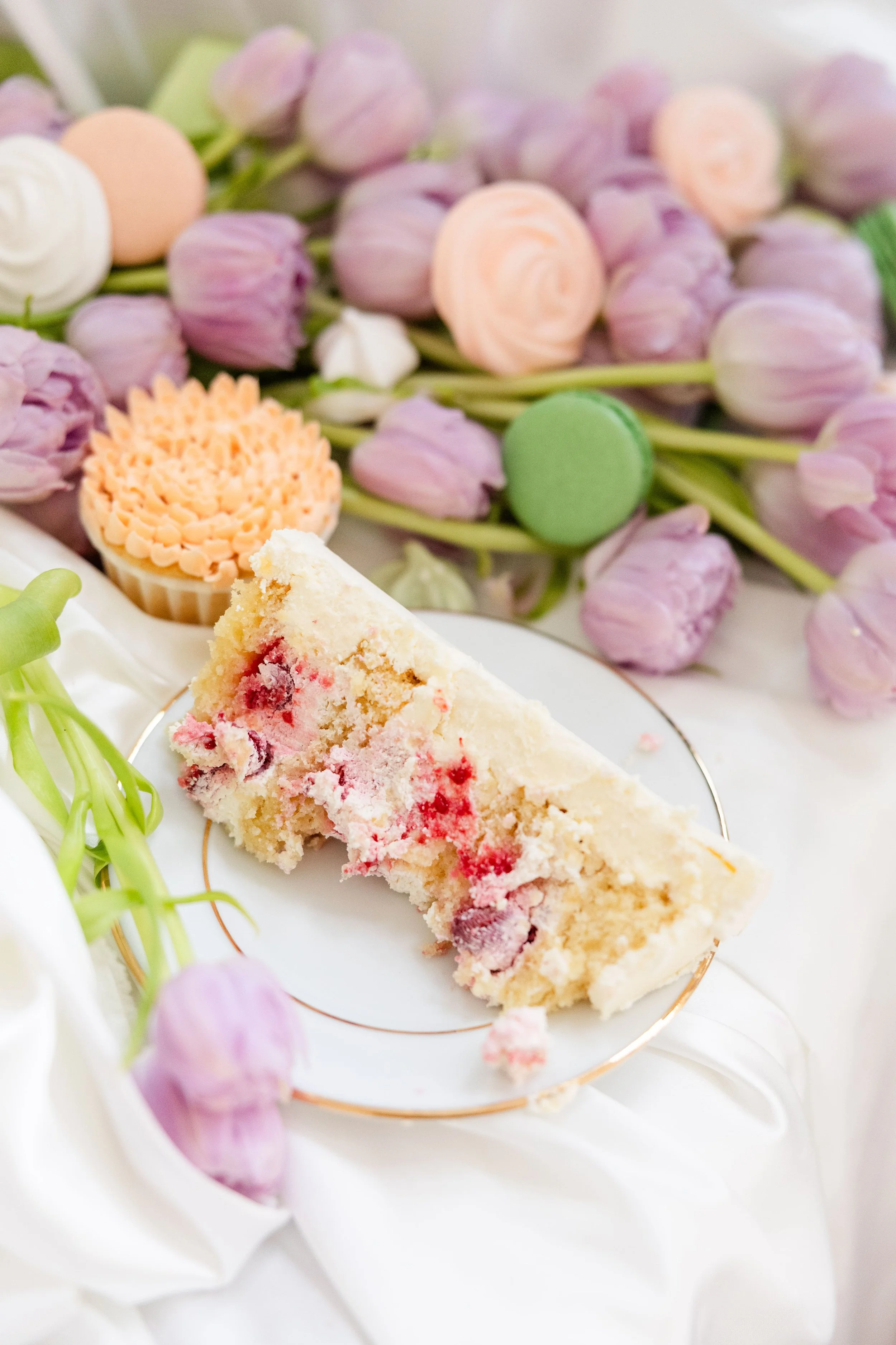 Part de gâteau de mariage aux fruits rouges présentée sur une table décorée de tulipes mauves, macarons et mignardises pastel.