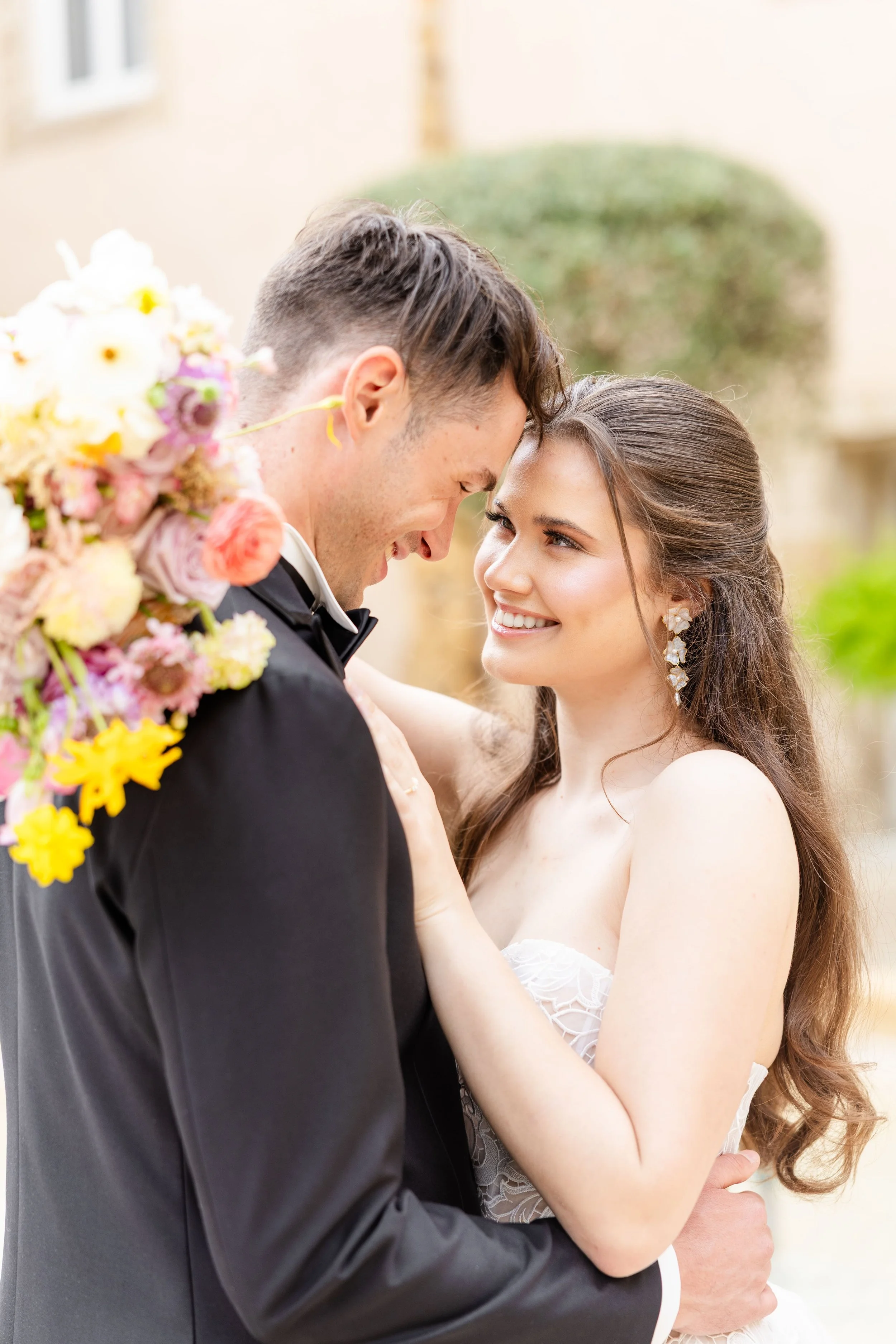 Portrait rapproché romantique des mariés s'enlaçant, la mariée tenant un bouquet de fleurs poudrées aux teintes violettes et jaunes pastel.