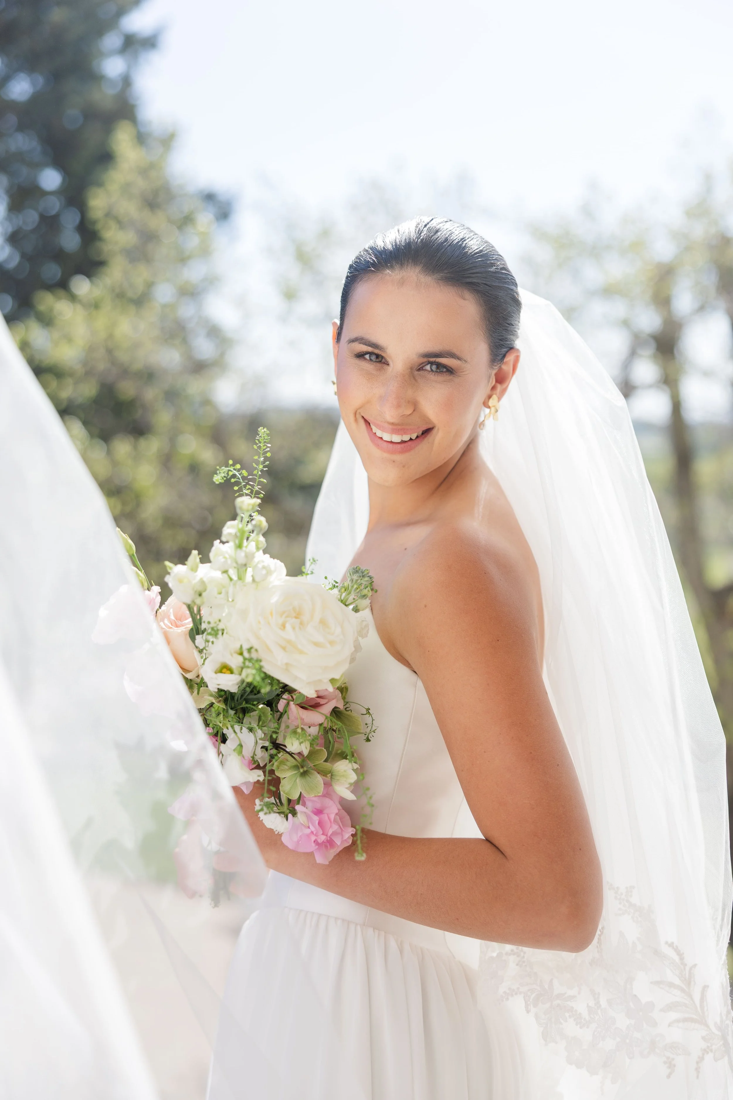 Portrait lumineux de la mariée souriante, portant un voile délicat en dentelle et tenant son bouquet de fleurs fraîches en extérieur.