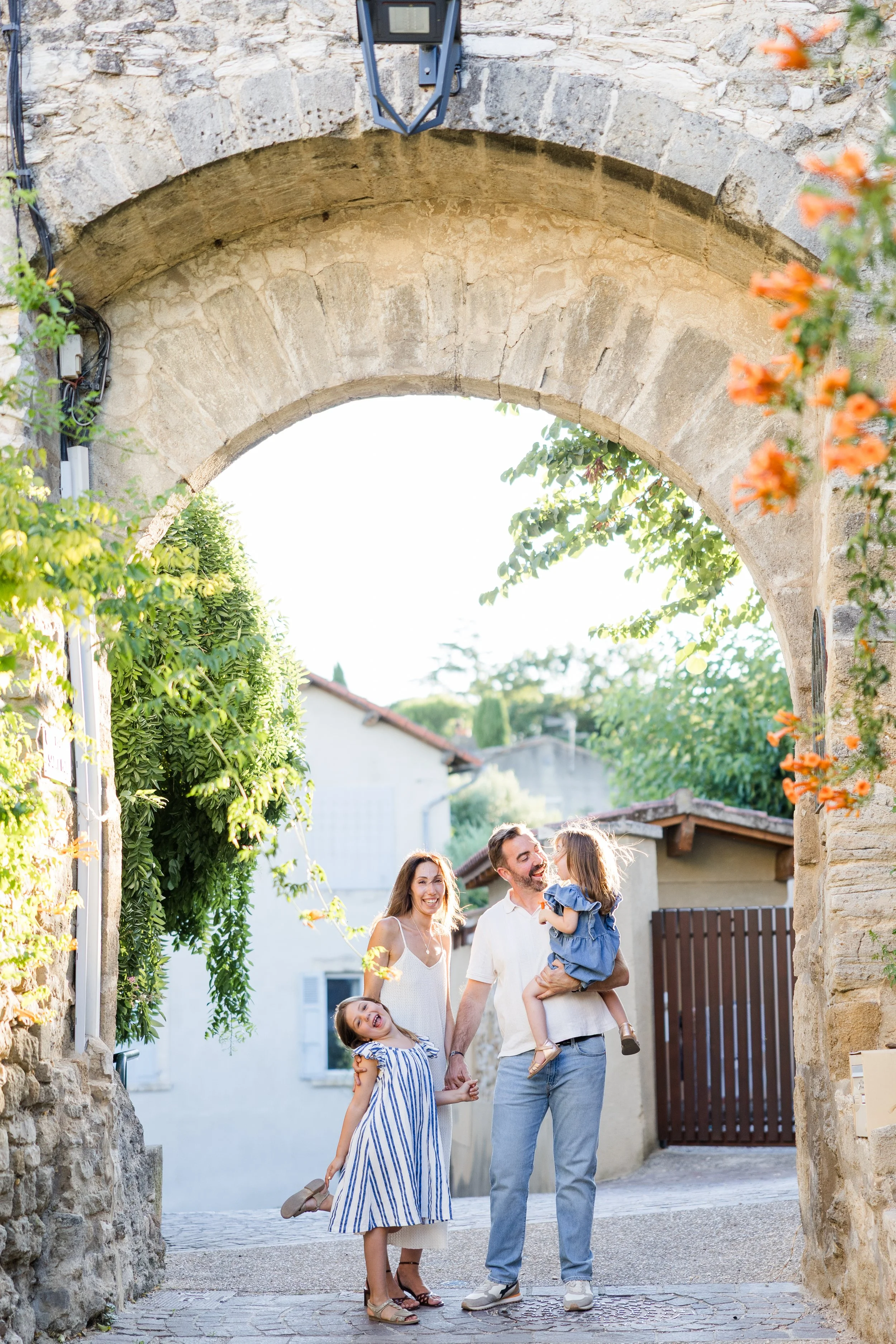 Une famille de quatre personnes posant joyeusement sous une arche ancienne en pierre, avec une maison en pierre et de la verdure en arrière-plan.