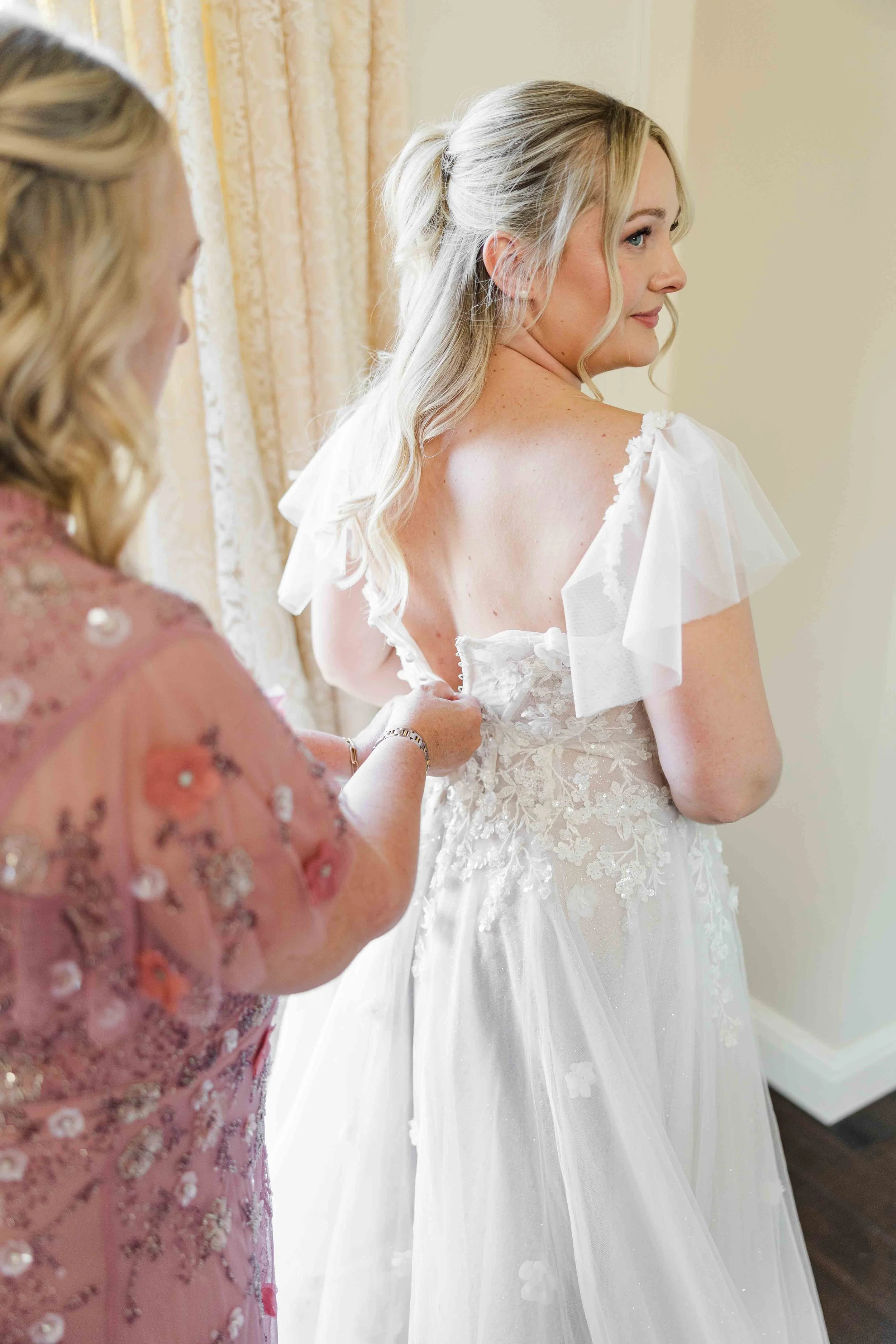 Moment complice des préparatifs où une proche aide à fermer le dos nu en dentelle et manches volantées de la robe de mariée.