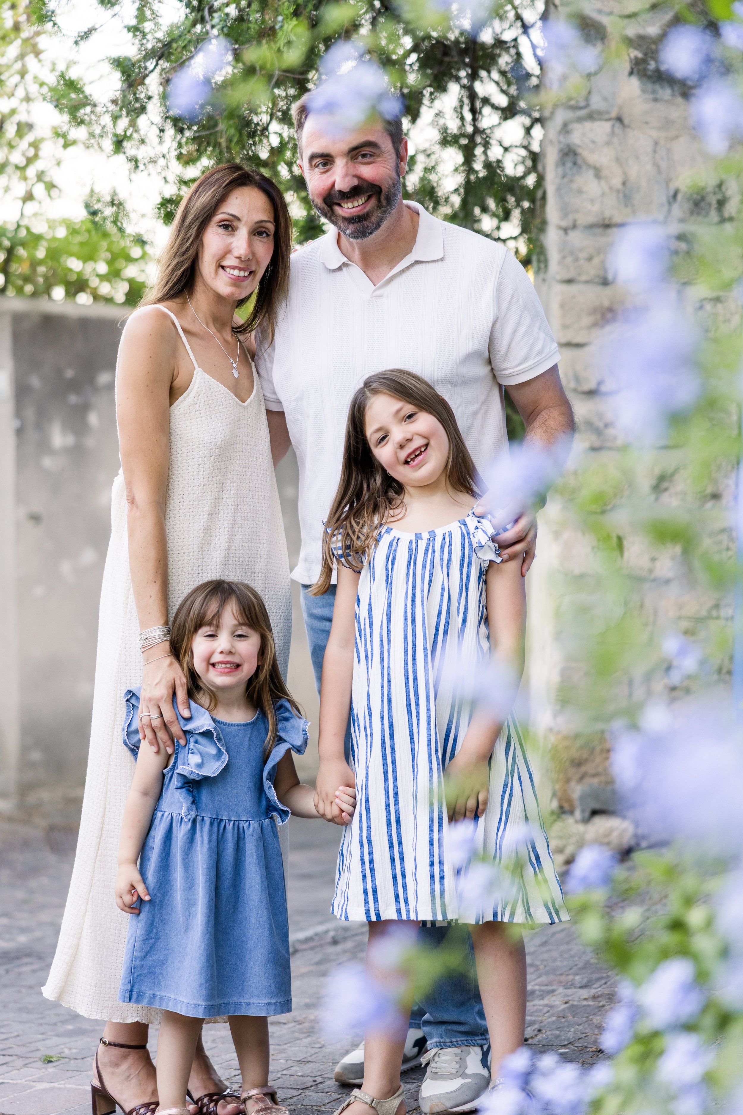 Une famille souriante de quatre personnes, vêtue de blanc et de rayures bleues, posant ensemble au premier plan, encadrée par de délicates fleurs violettes en Provence.