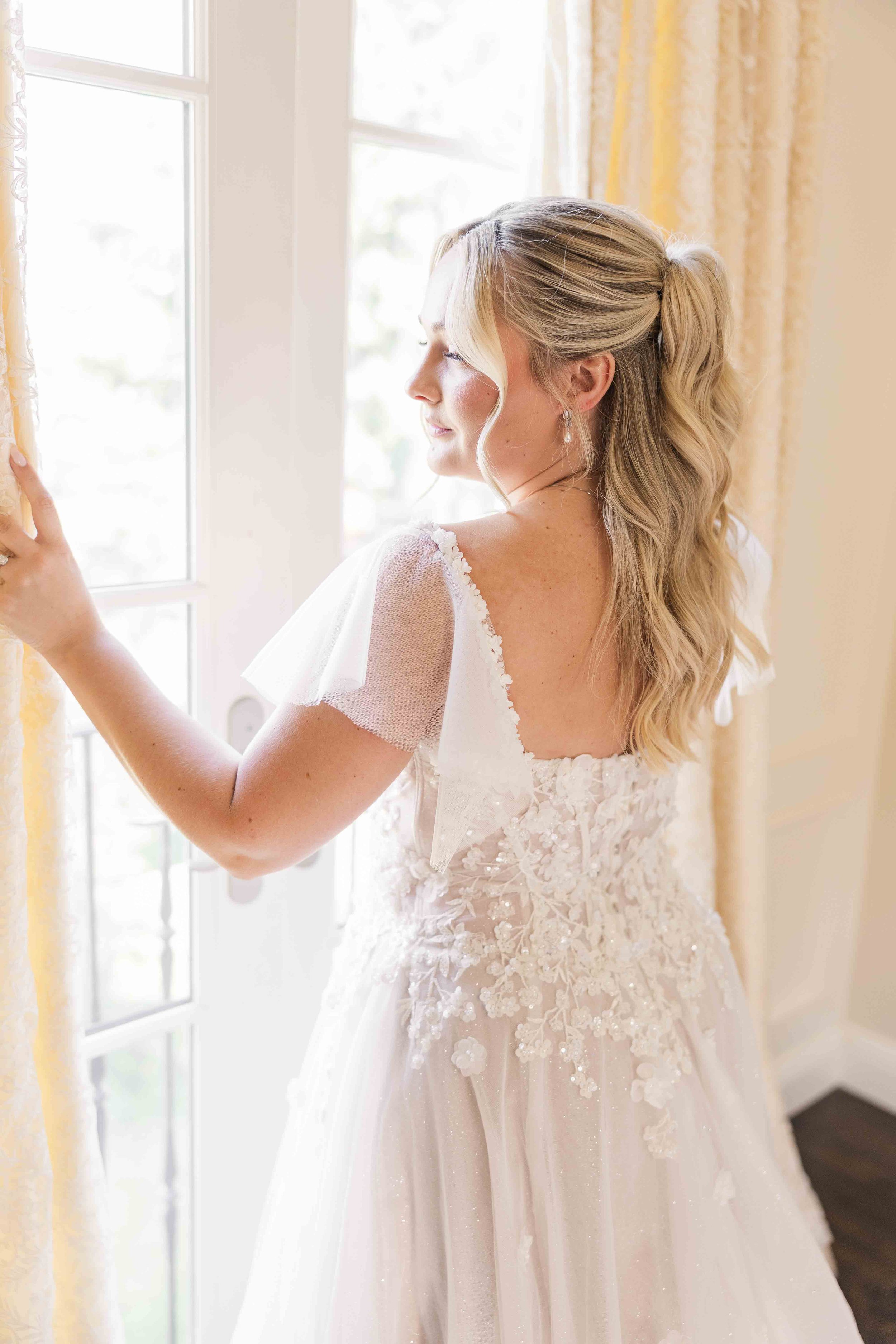 Portrait lumineux de la mariée coiffée d'une demi-queue avec des ondulations, regardant par la fenêtre dans sa robe à détails floraux et manches en tulle.