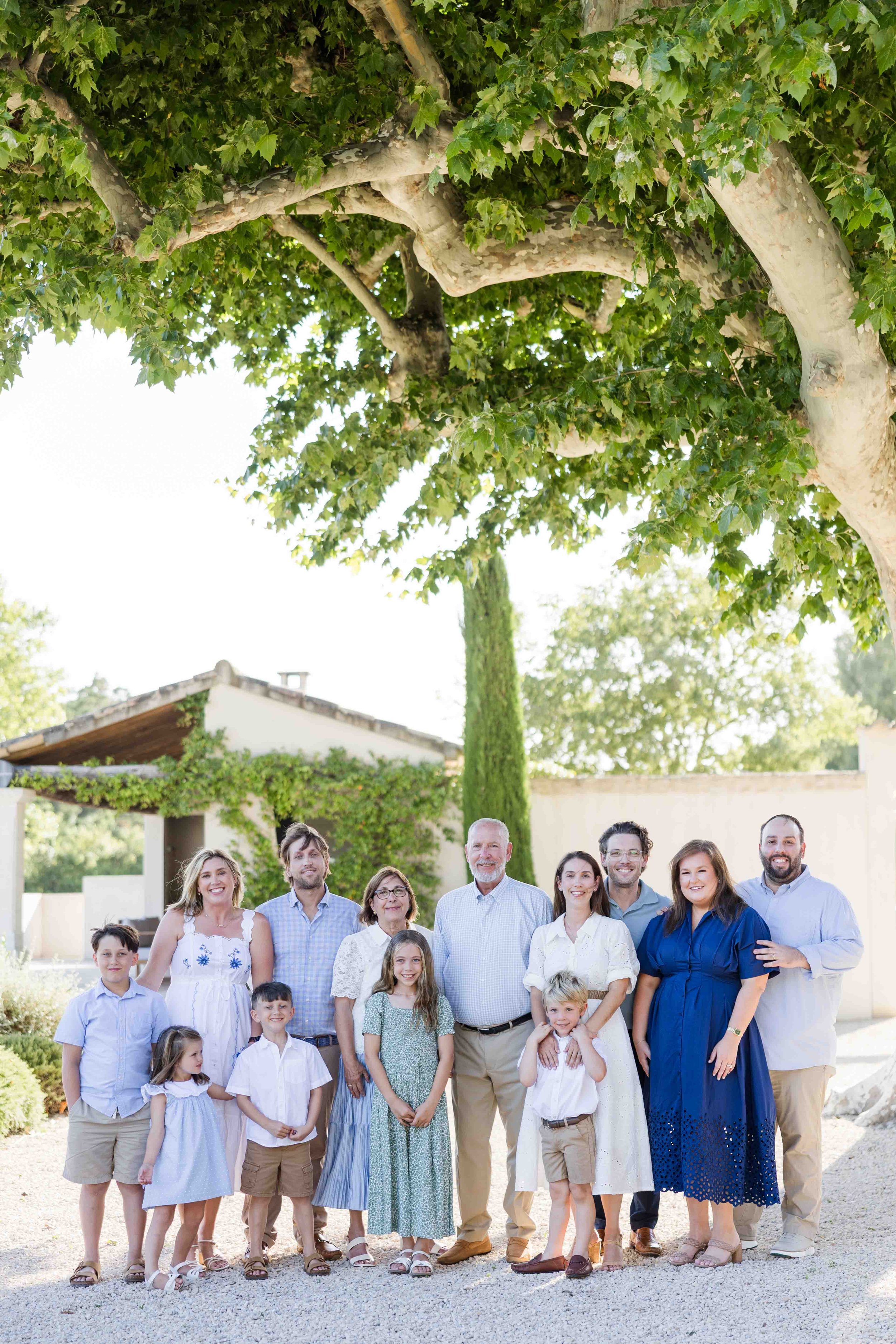 Photo de groupe d'une grande famille intergénérationnelle posant à l'ombre d'un grand platane dans la cour d'un domaine provençal.