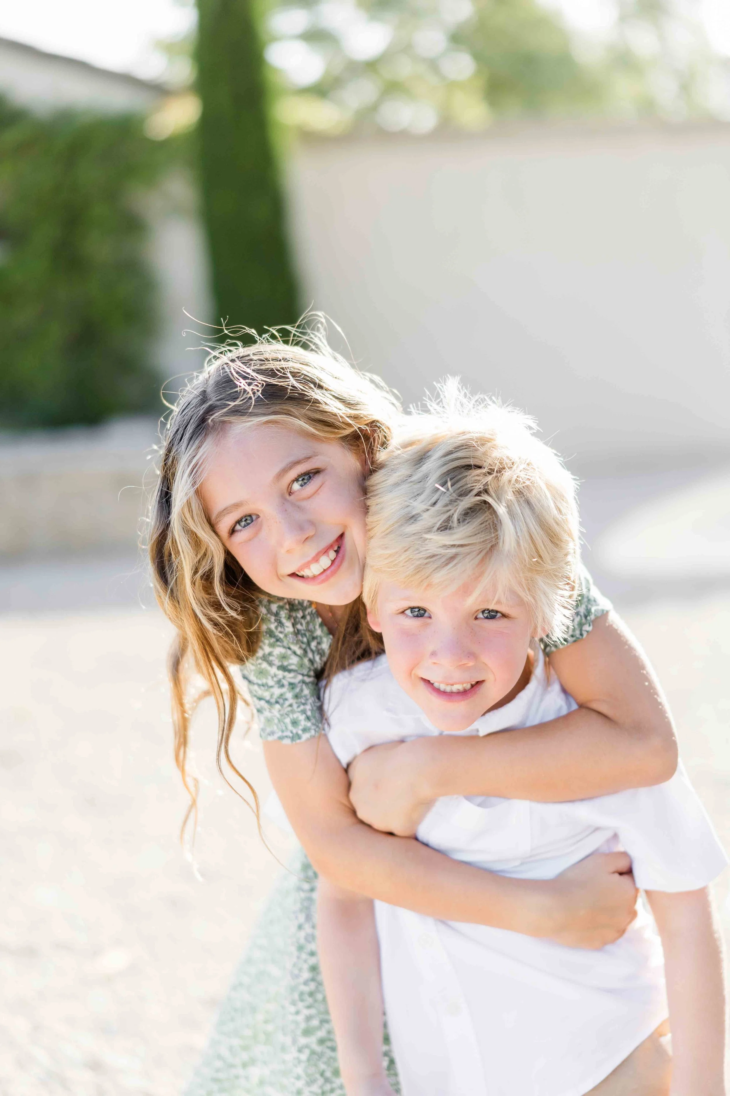 Portrait plein de tendresse d'une grande sœur enlacant son petit frère de dos, tous deux souriant à l'objectif au soleil.