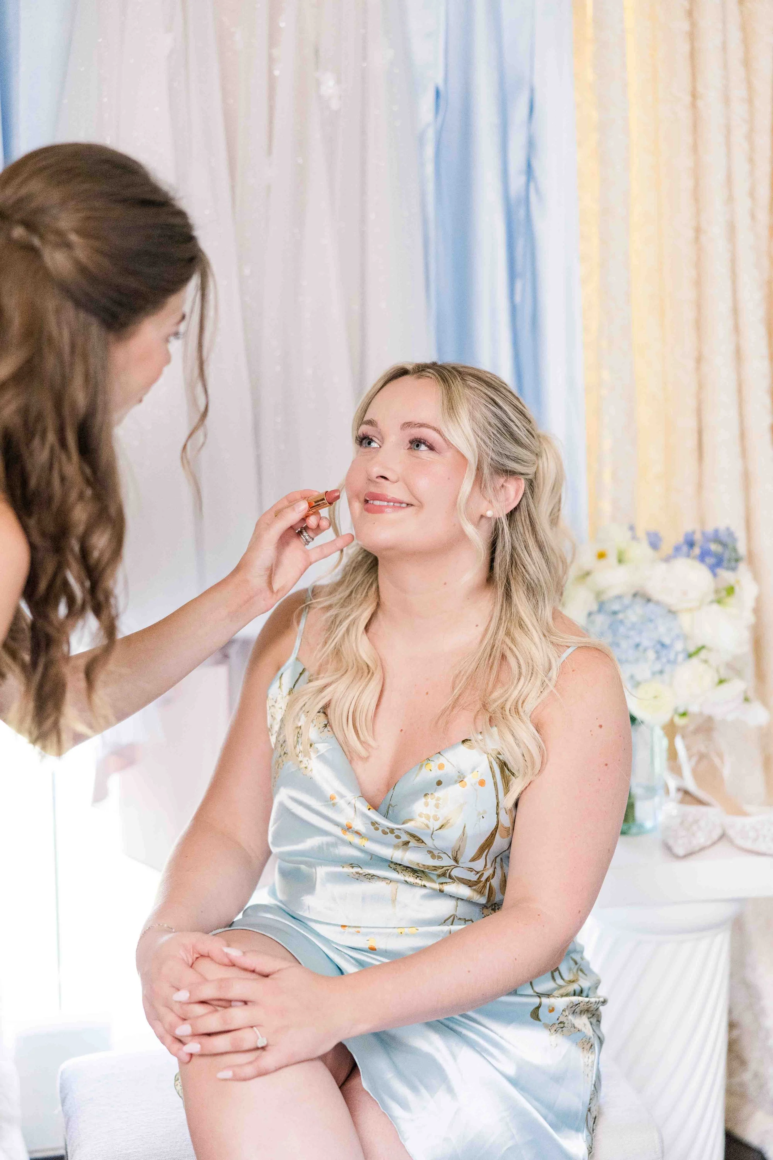 Séance de maquillage de la mariée vêtue d'un peignoir en soie bleue à motifs floraux, entourée de légers voiles colorés.