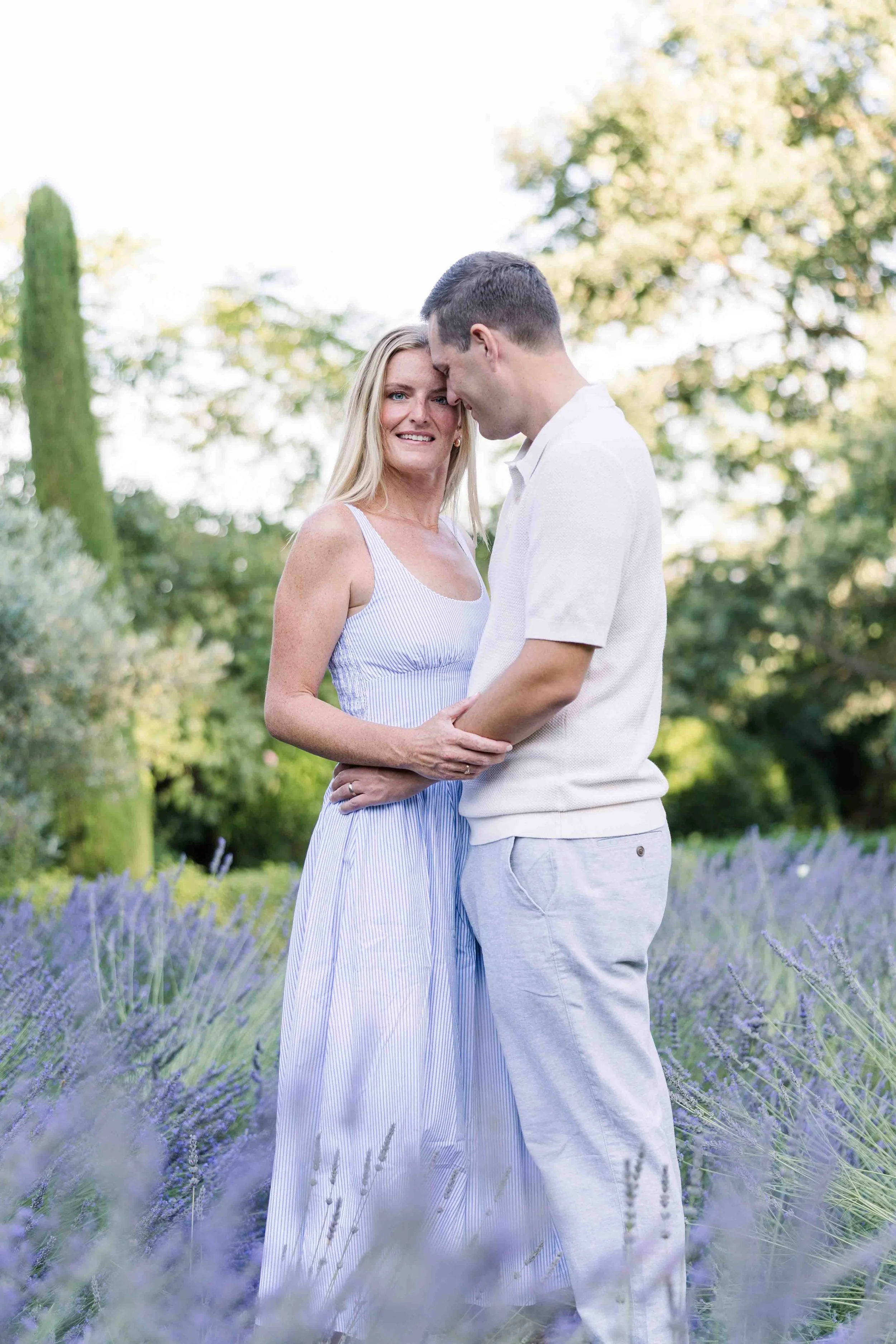 Un couple enlacé tendrement au premier plan d'un champ de lavande en Provence, devant une allée de cyprès. La femme porte une robe bleu clair, l'homme une chemise beige.