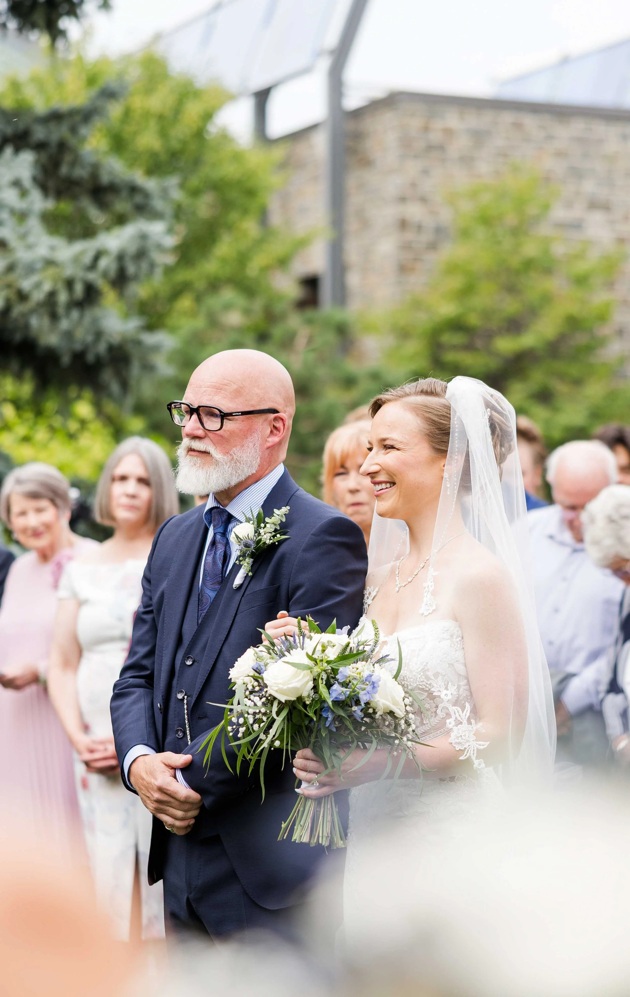 La mariée au bras de son père portant un costume bleu marine et des lunettes lors de l'entrée de cérémonie en extérieur.