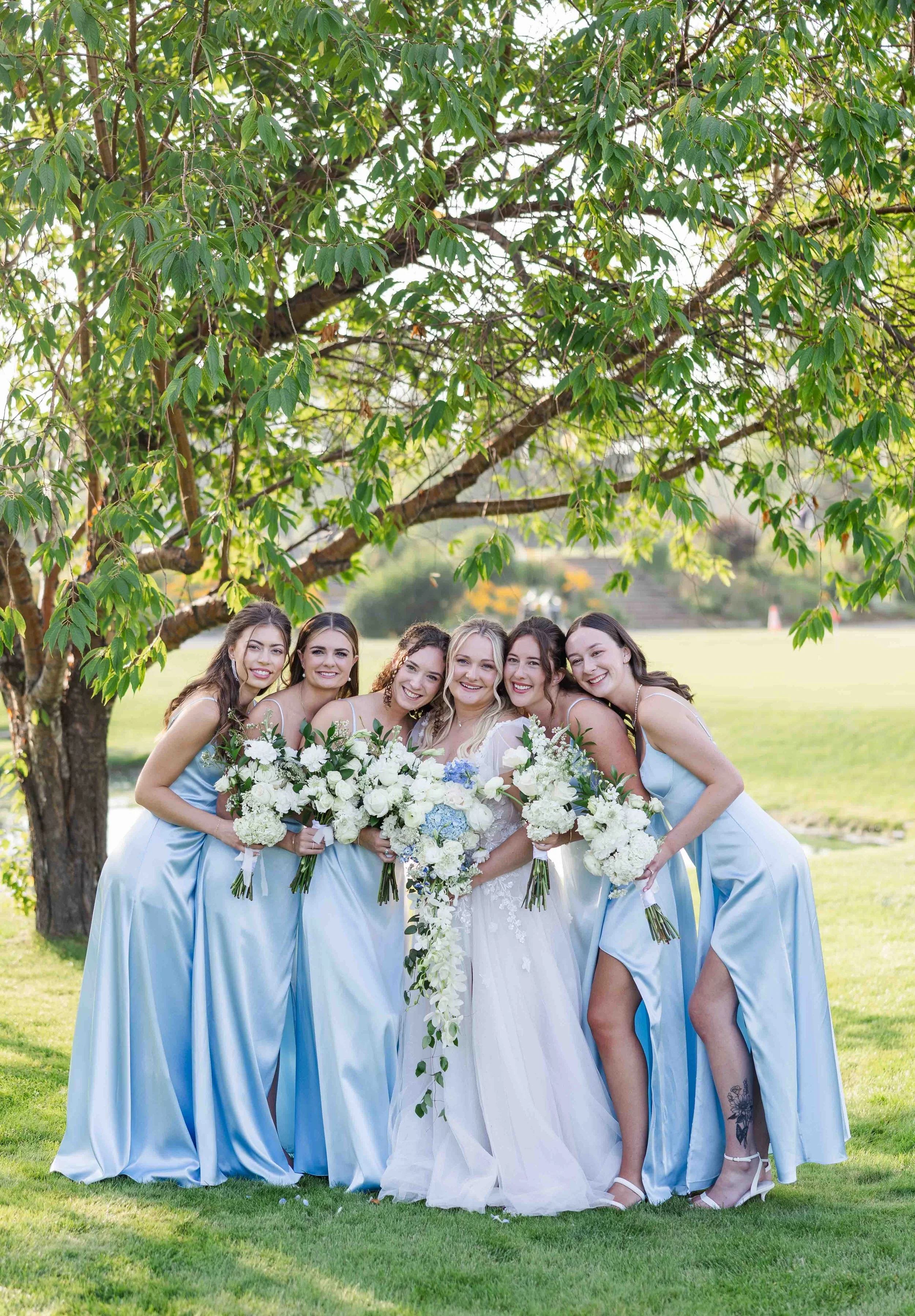 La mariée et ses demoiselles d'honneur souriantes posant ensemble en extérieur sous les arbres avec leurs bouquets de fleurs blanches et feuillage.