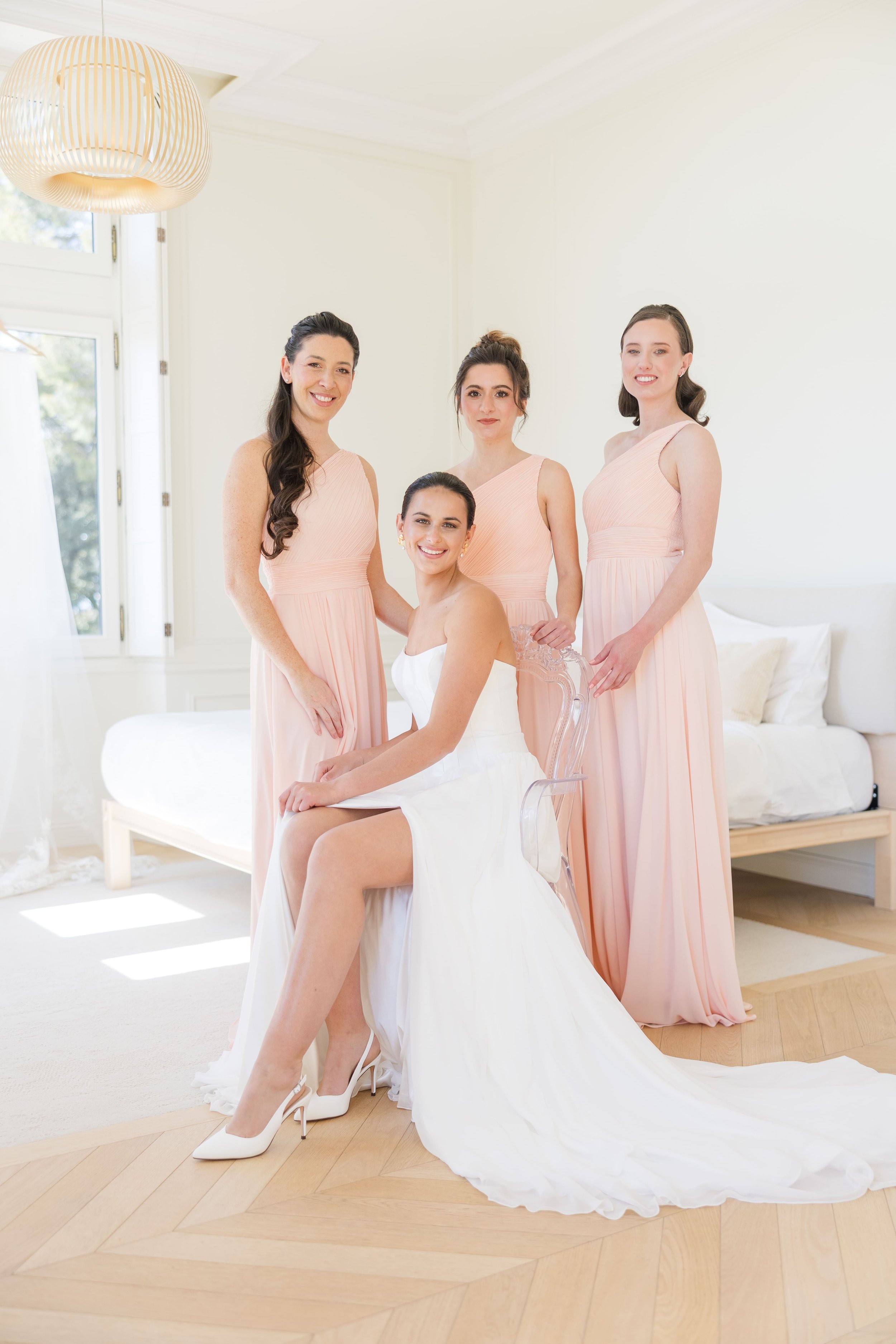 La mariée assise entourée de ses trois demoiselles d'honneur vêtues de robes longues couleur pêche dans une suite lumineuse.