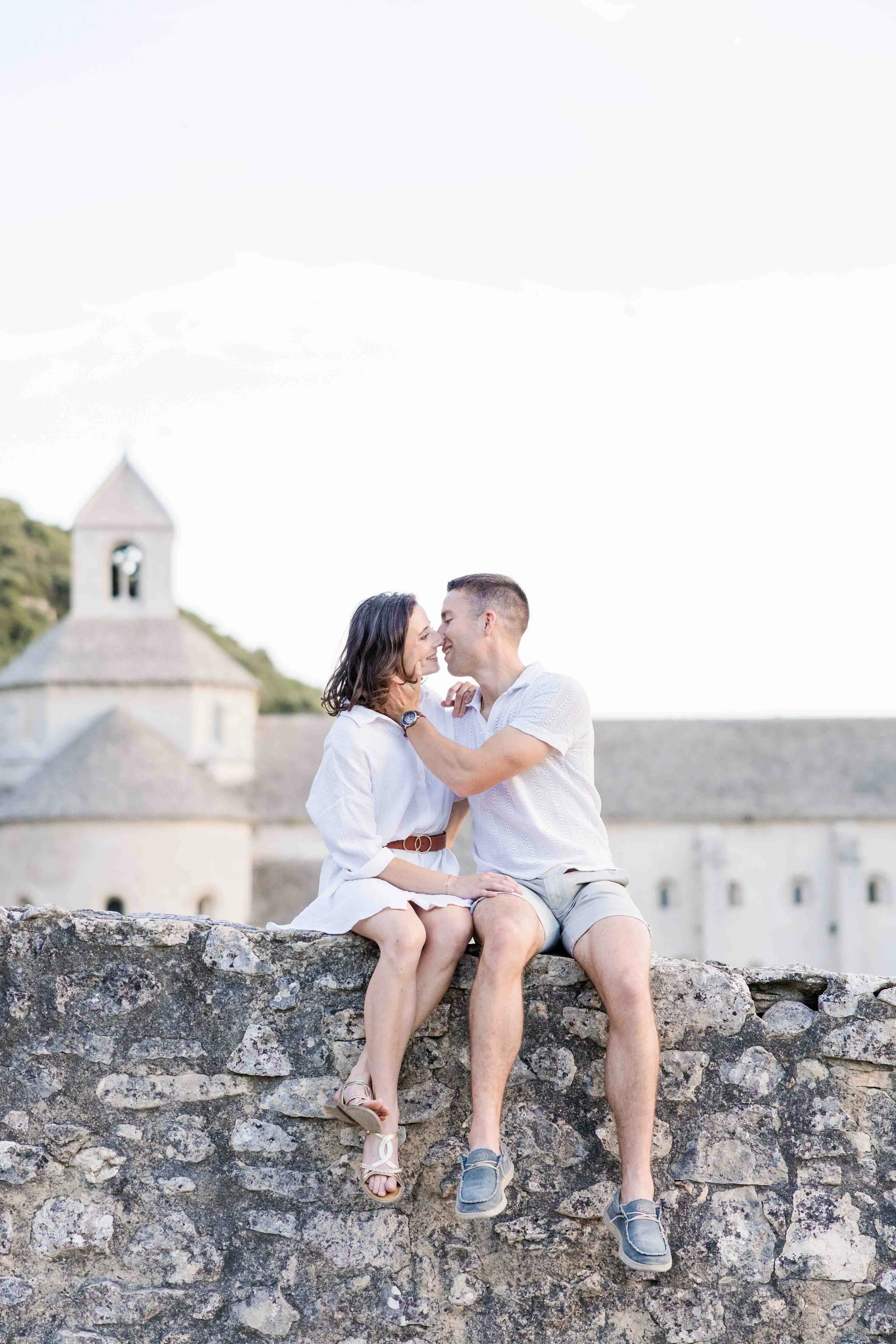 Un couple assis sur un mur en pierre ancienne, échangeant un regard tendre avec le clocher de l'Abbaye de Sénanque en fond.