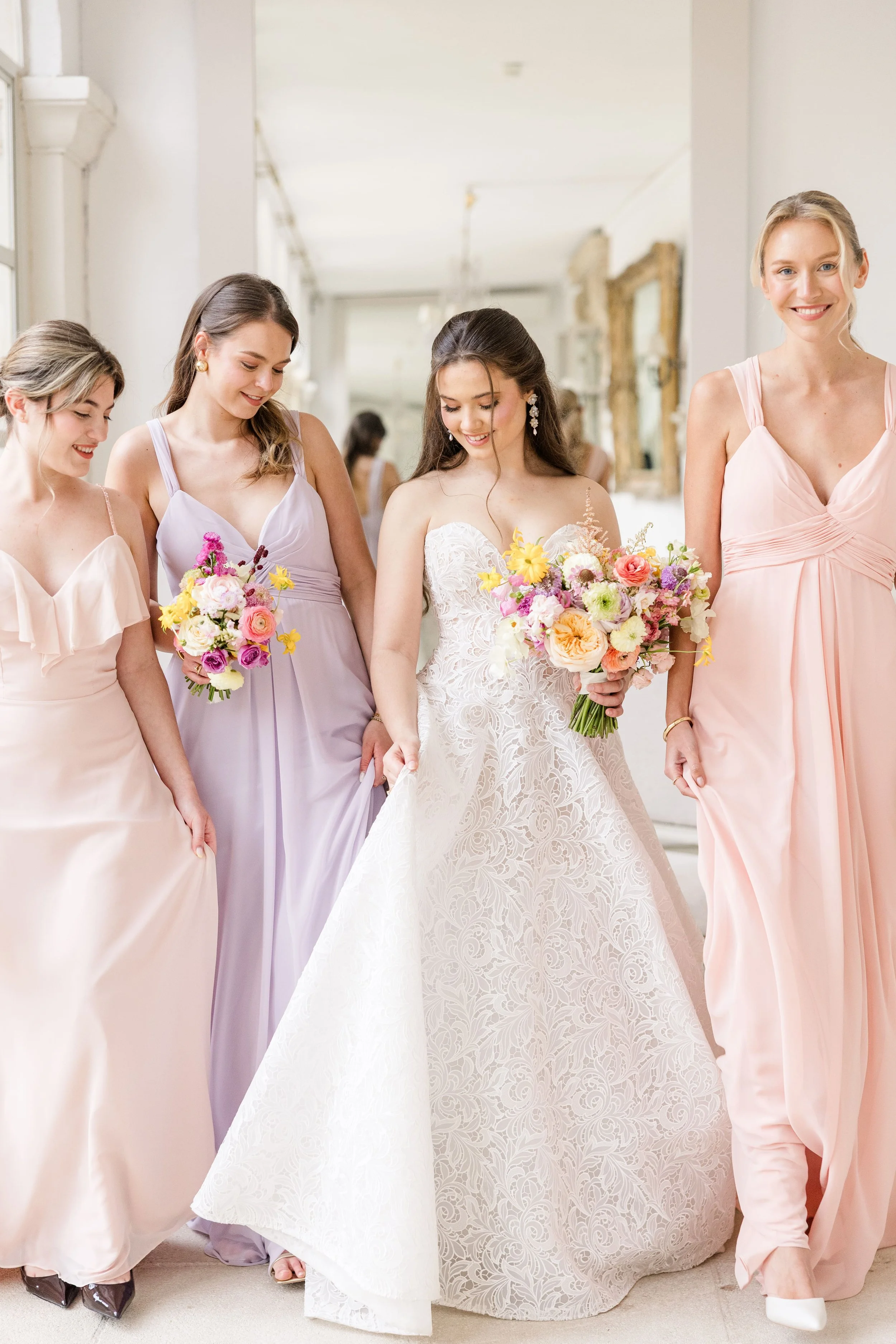 La mariée marchant aux côtés de ses demoiselles d'honneur vêtues de robes pastel roses et violettes dans les salons d'un château provençal.