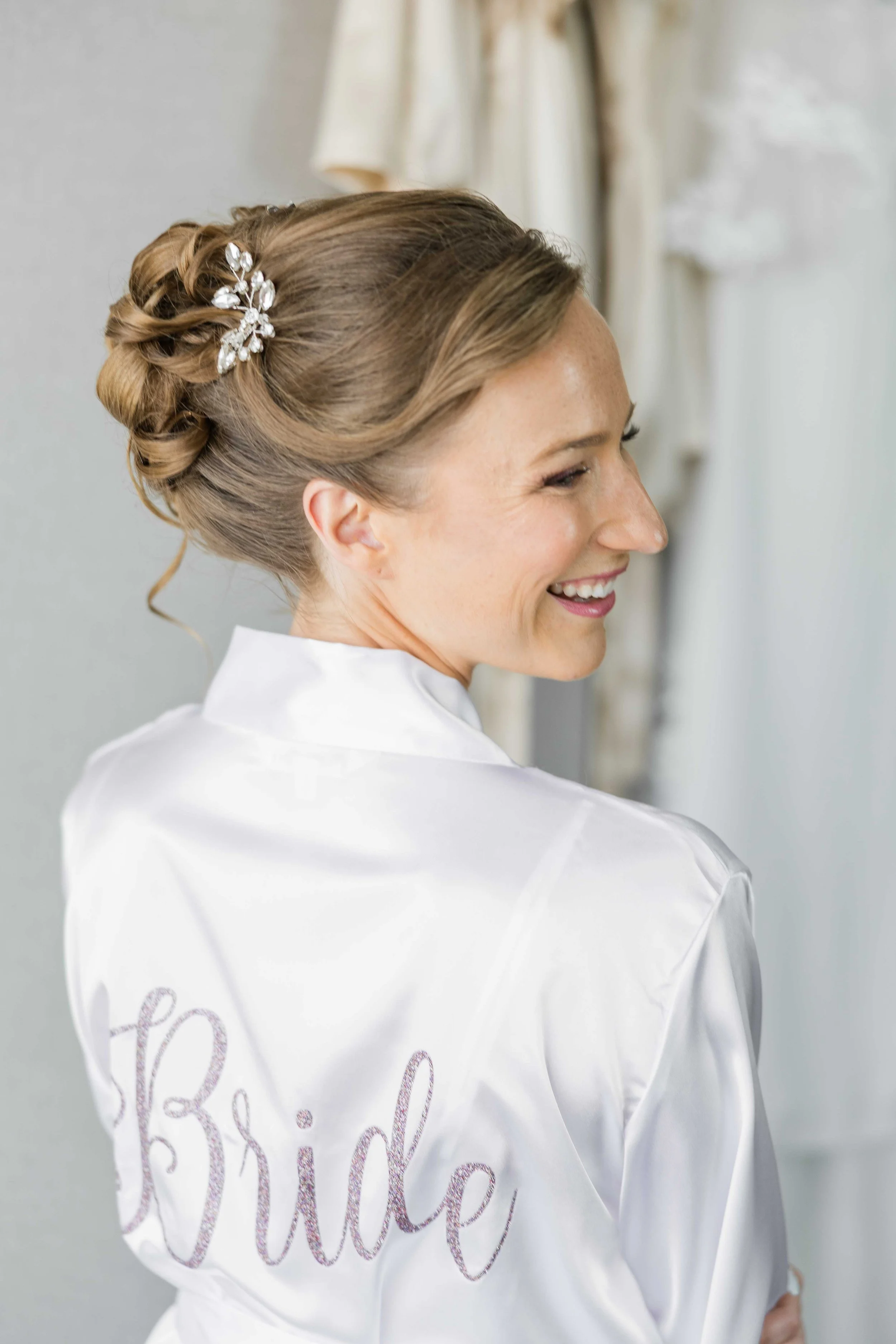 La mariée de dos en peignoir en soie blanche brodé Bride, dévoilant son chignon d'un style élégant agrémenté d'un bijou de tête.