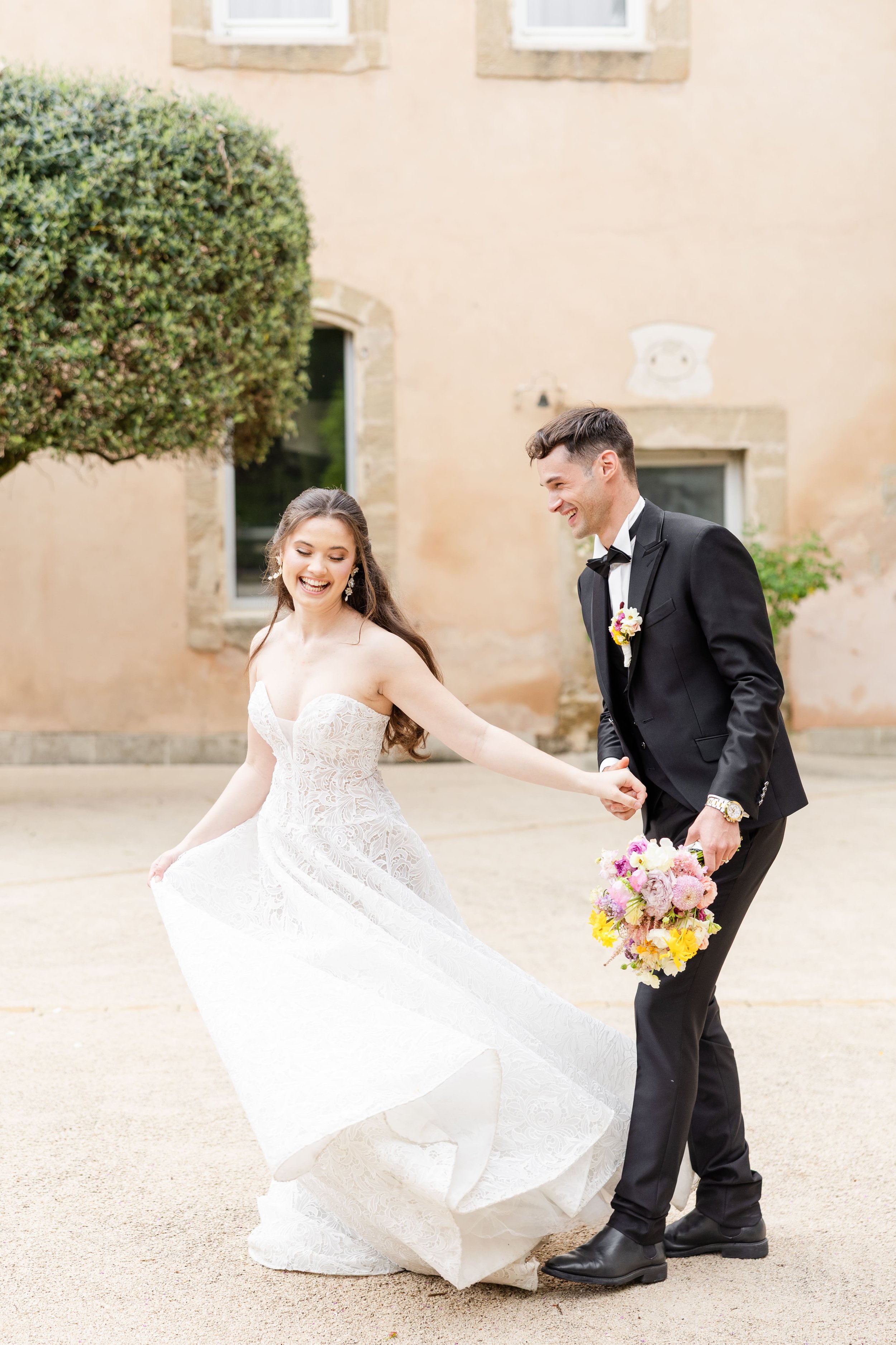Mariée en robe princesse dentelle et marié en costume noir dansant joyeusement dans la cour d'un château élégant en Provence.