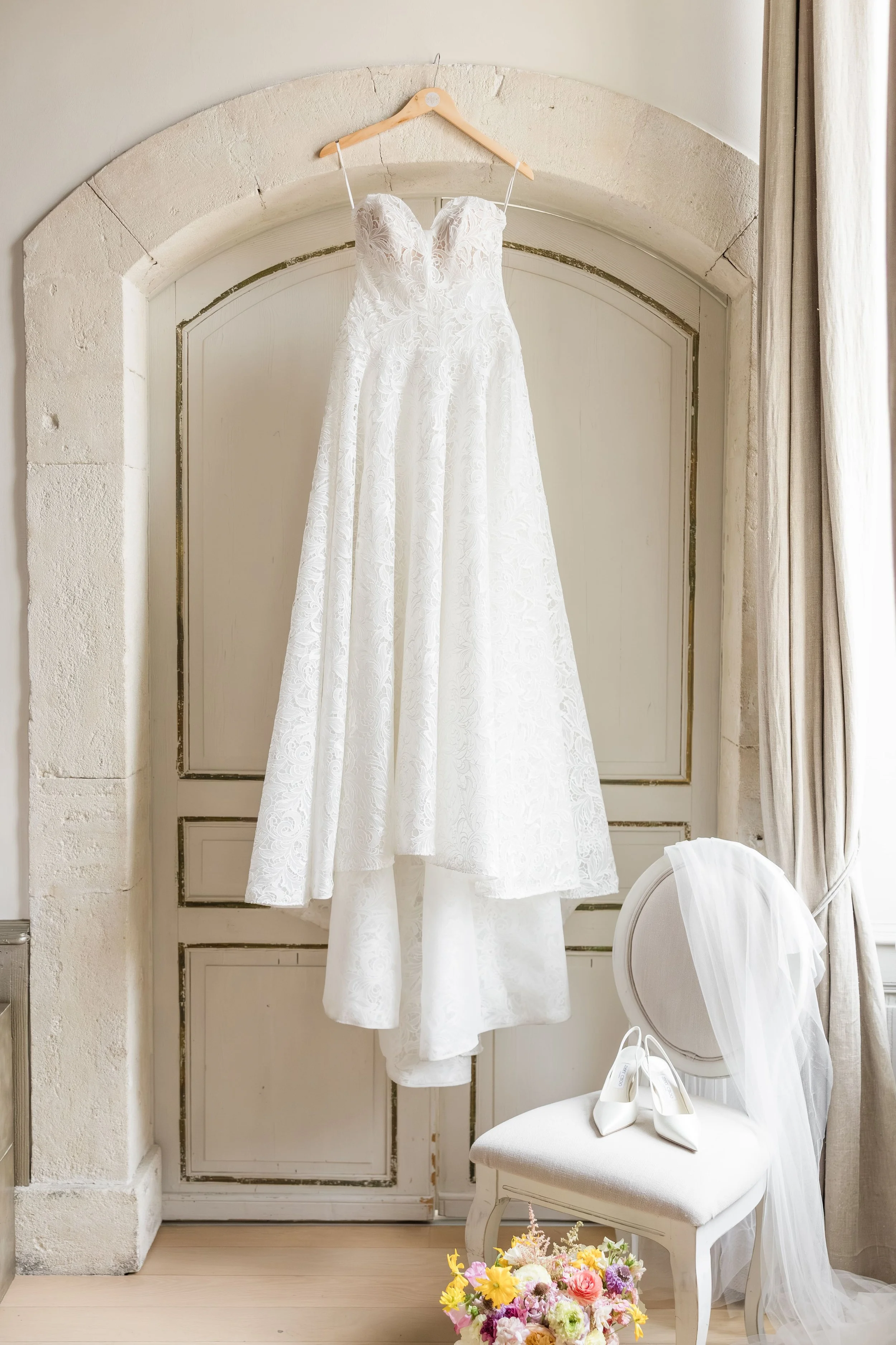 Robe de mariée bustier en dentelle suspendue devant une porte en pierre sculptée, détails des préparatifs dans un domaine de mariage en Provence.