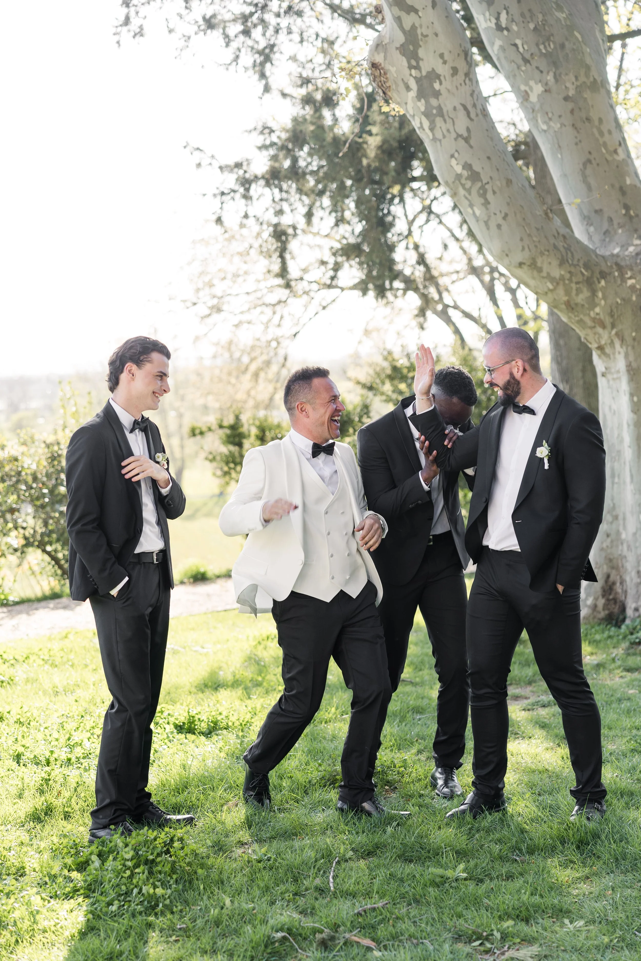 Le marié et ses garçons d'honneur en smokings élégants partageant un moment de rire et de complicité dans le parc d'un domaine.