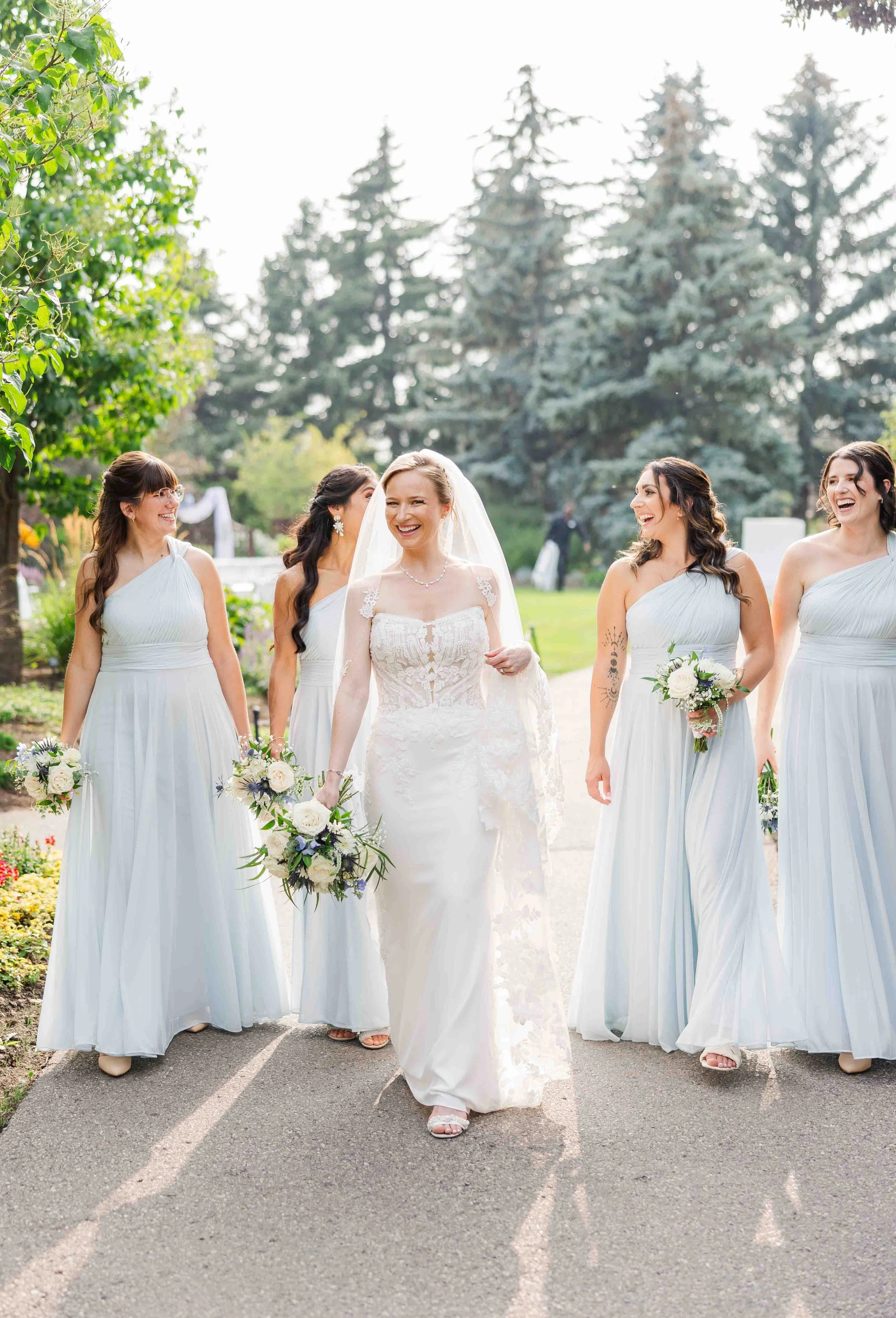 La mariée marchant dans une allée bordée d'arbres, entourée de son cortège de demoiselles d'honneur en robes bleu pastel.