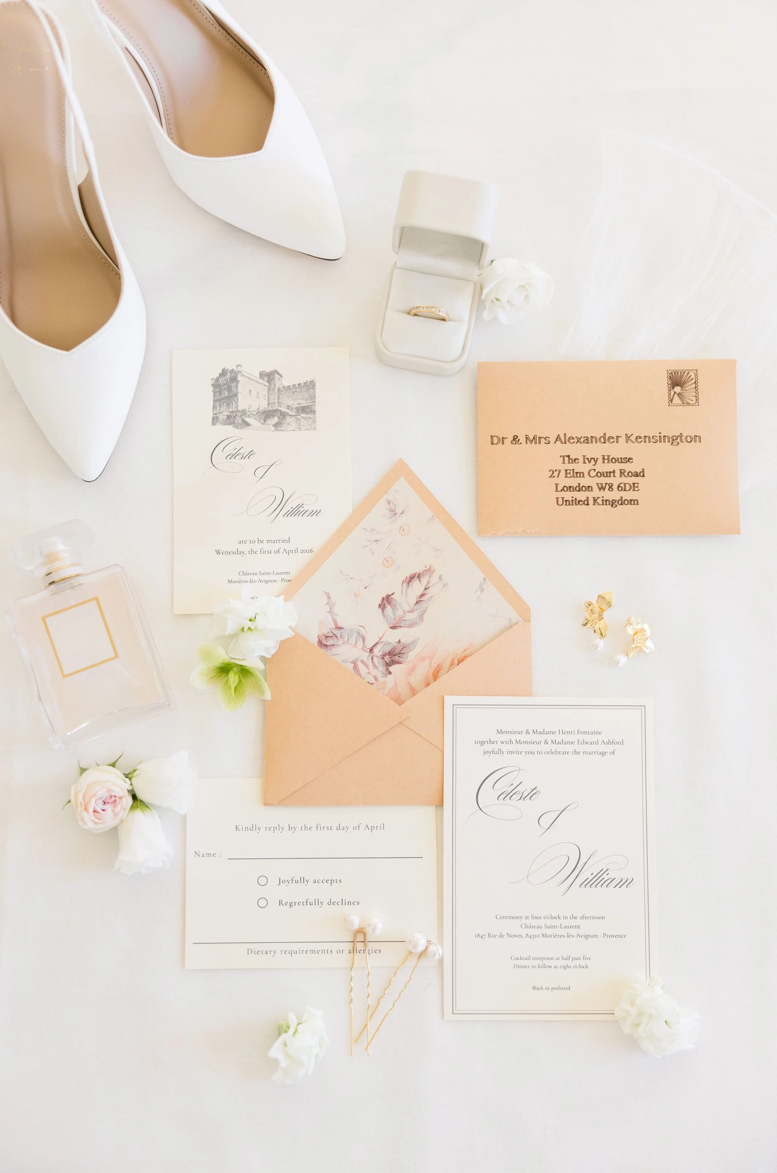 Flatlay de papeterie de mariage haut de gamme avec faire-part dorés, chaussures blanches à talons, flacon de parfum et alliances.