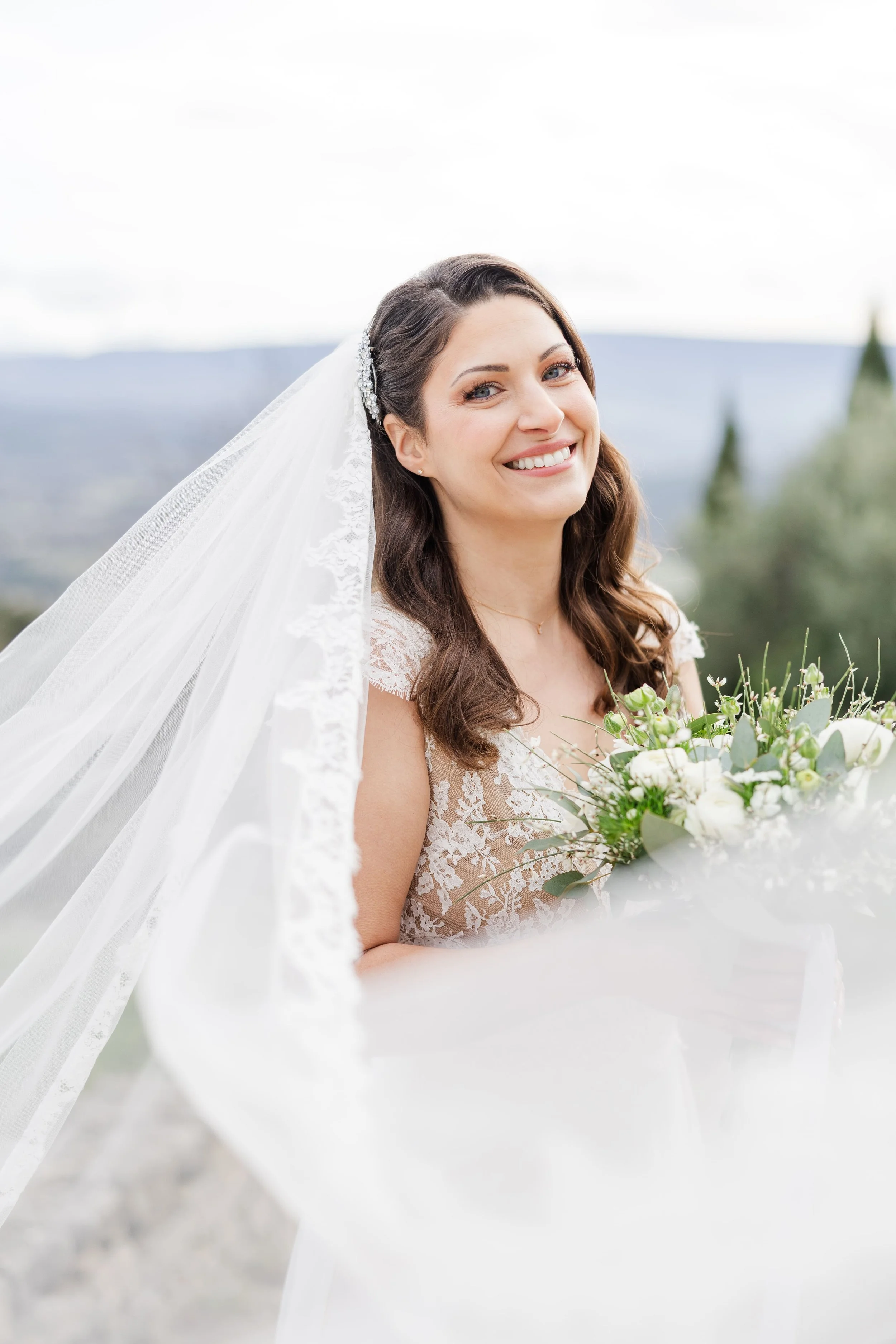 Portrait de la mariée souriante, son long voile en tulle volant au vent, sur fond de paysage provençal à Gordes.