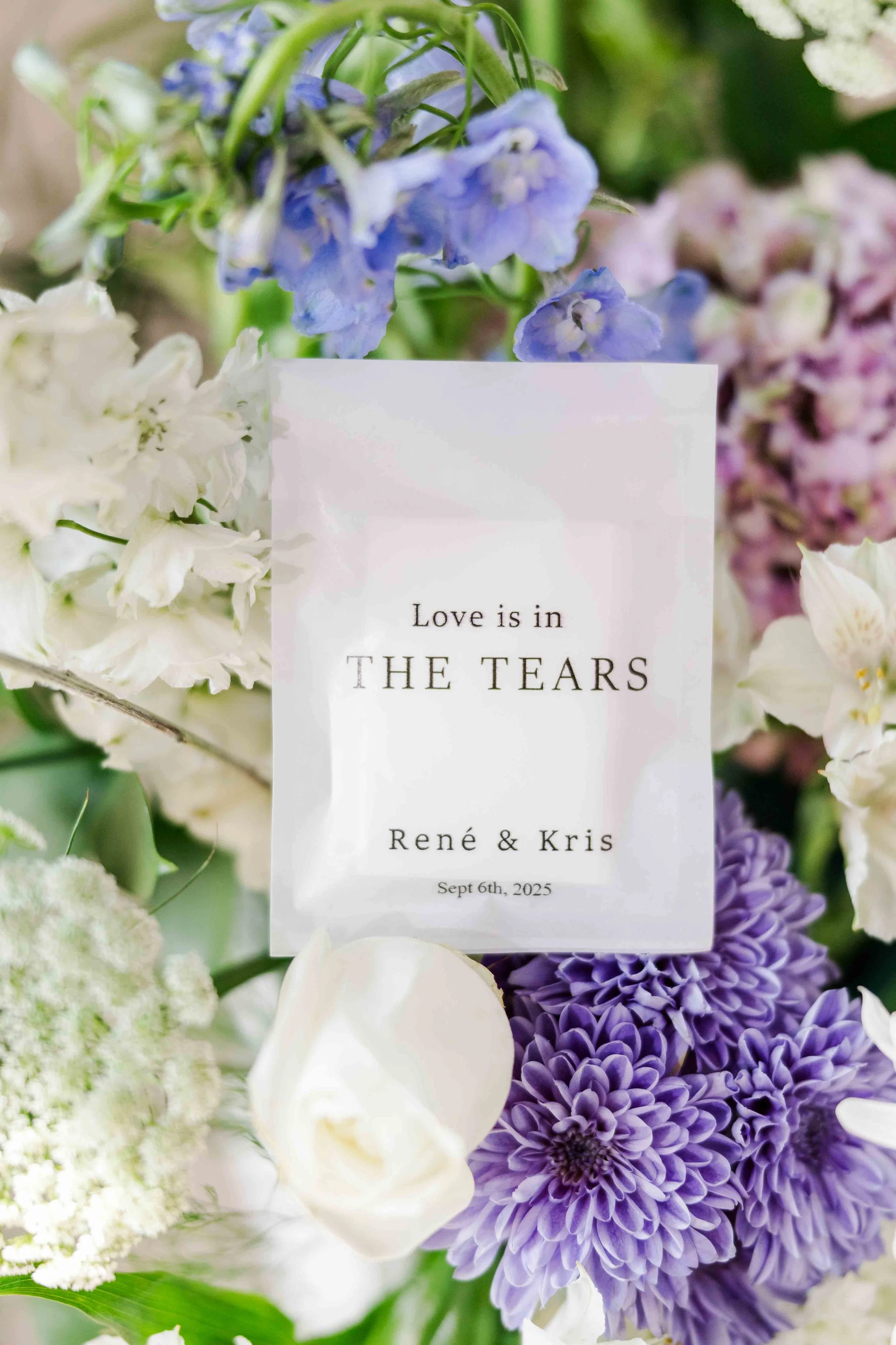 Flatlay de détails de mariage montrant un sachet personnalisé « For Your Happy Tears » entouré d'hortensias bleus, violets et de roses blanches.