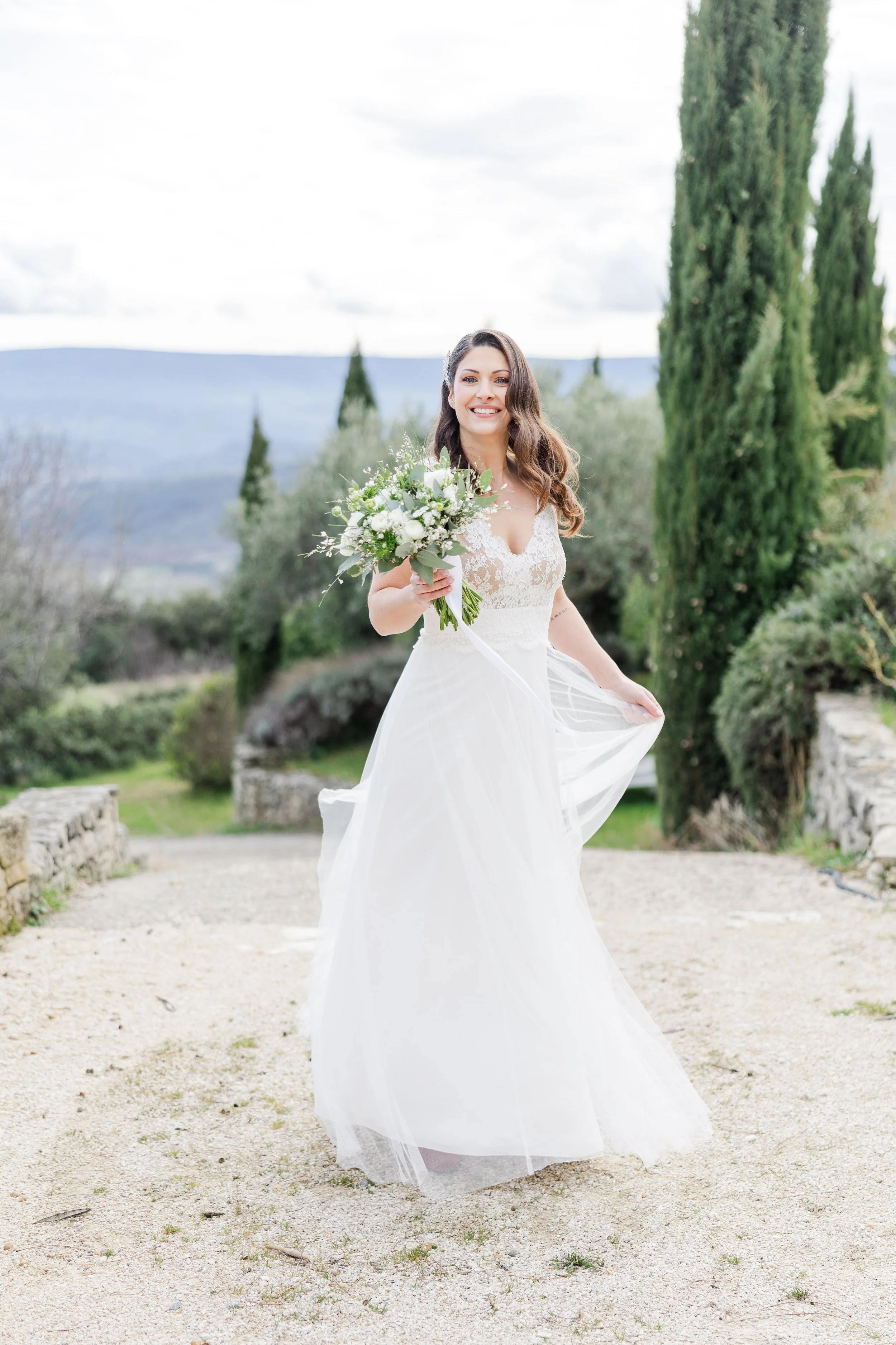 Portrait plein pied de la mariée en robe fluide bretelles, tenant son bouquet d'eucalyptus devant des cyprès en Provence.