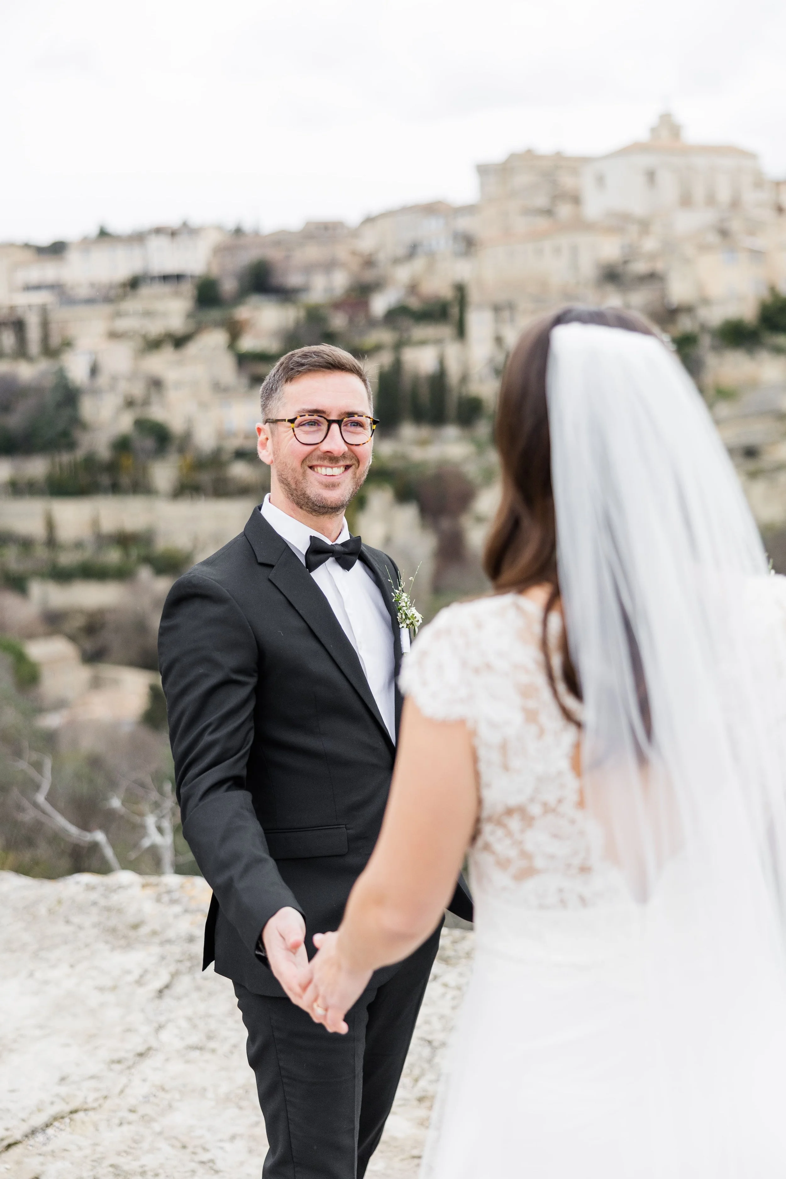 Le marié en smoking noir et la mariée de dos se tenant par la main face au panorama emblématique du village de Gordes.