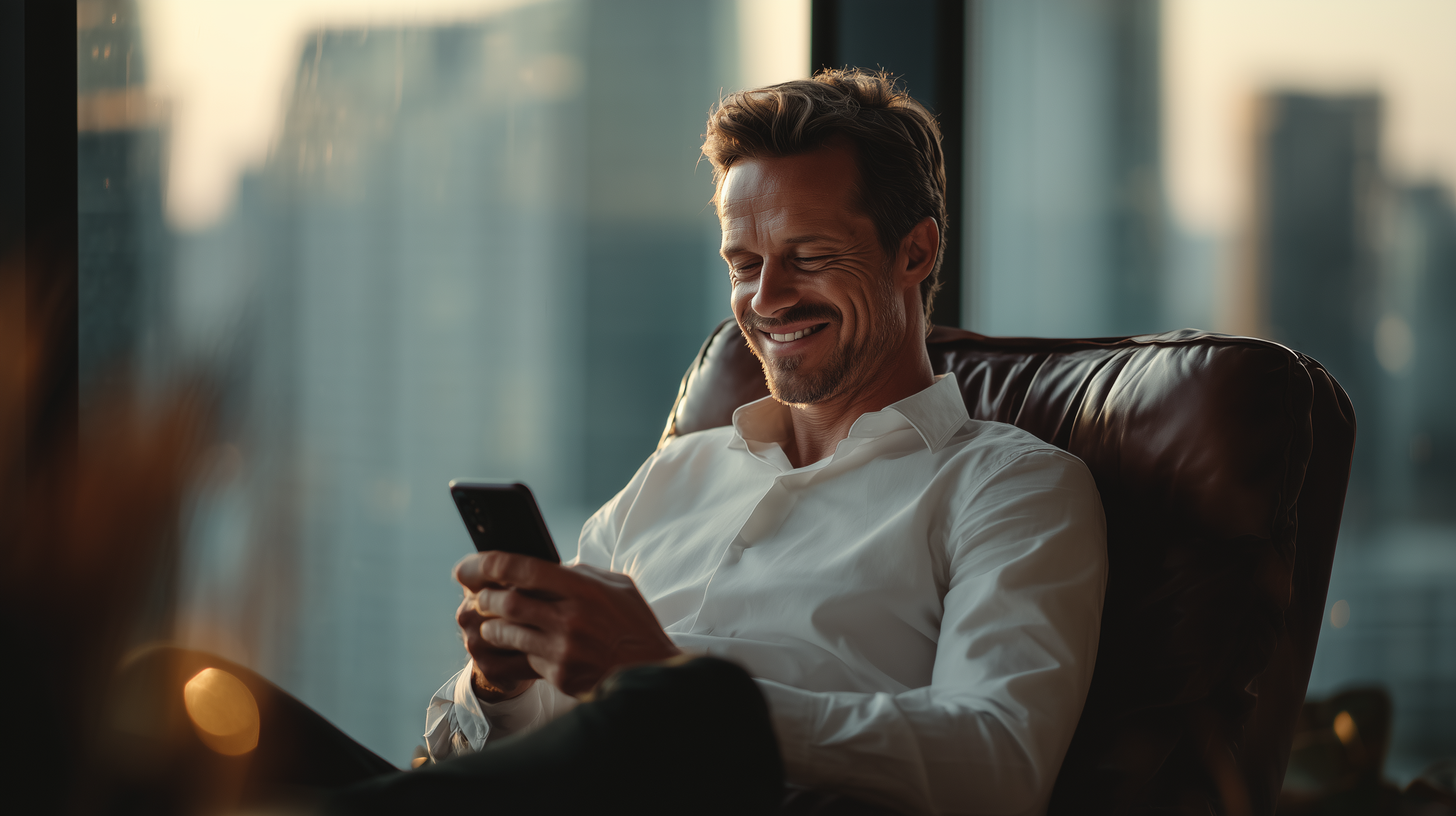 Homem sorridente sentado em uma poltrona de couro, usando camisa branca, olhando para o telefone móvel com uma vista de uma cidade ao fundo.