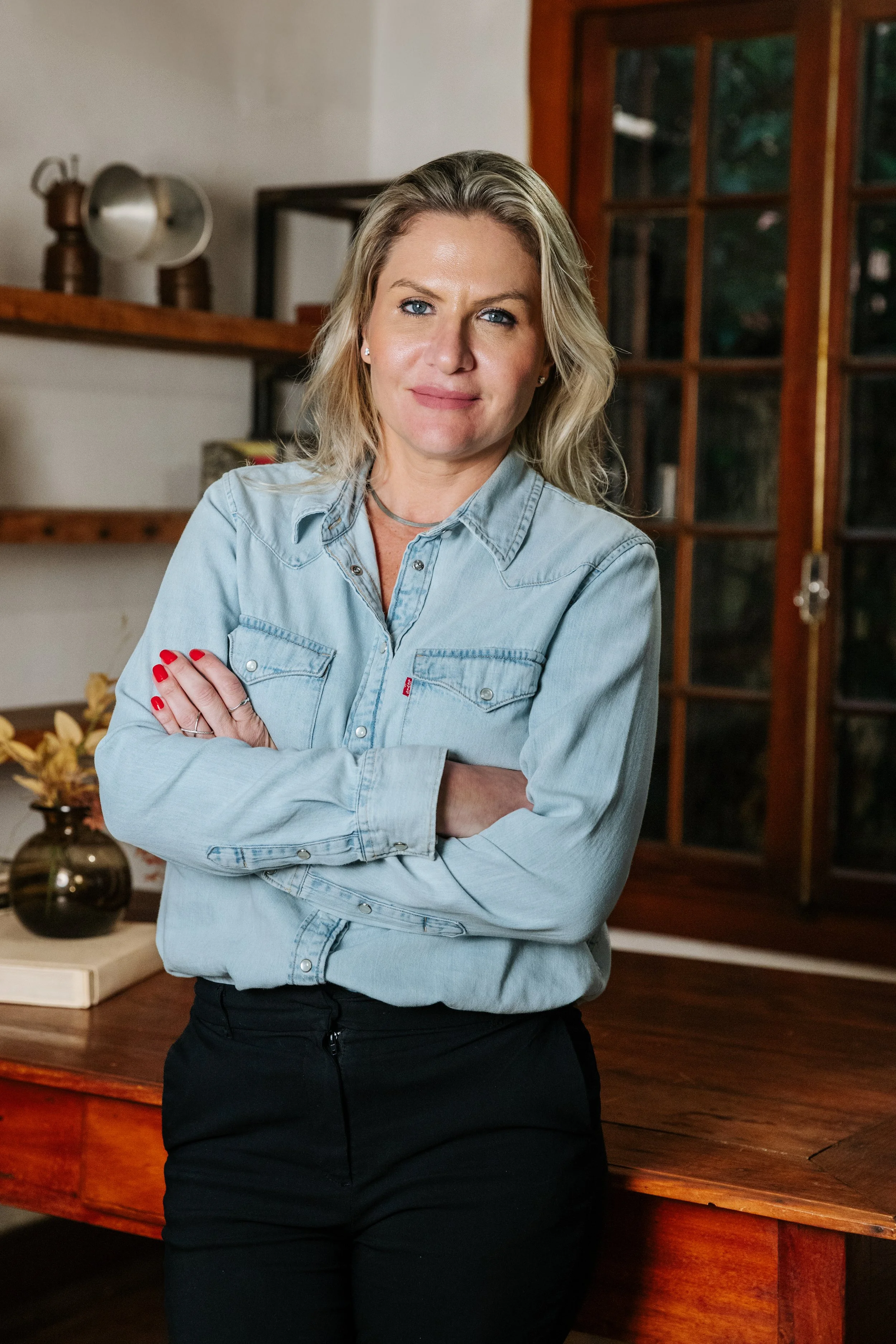 Mulher com cabelos loiros, usando blusa jeans azul de mangas longas e calça preta, cruzando os braços, em ambiente interno