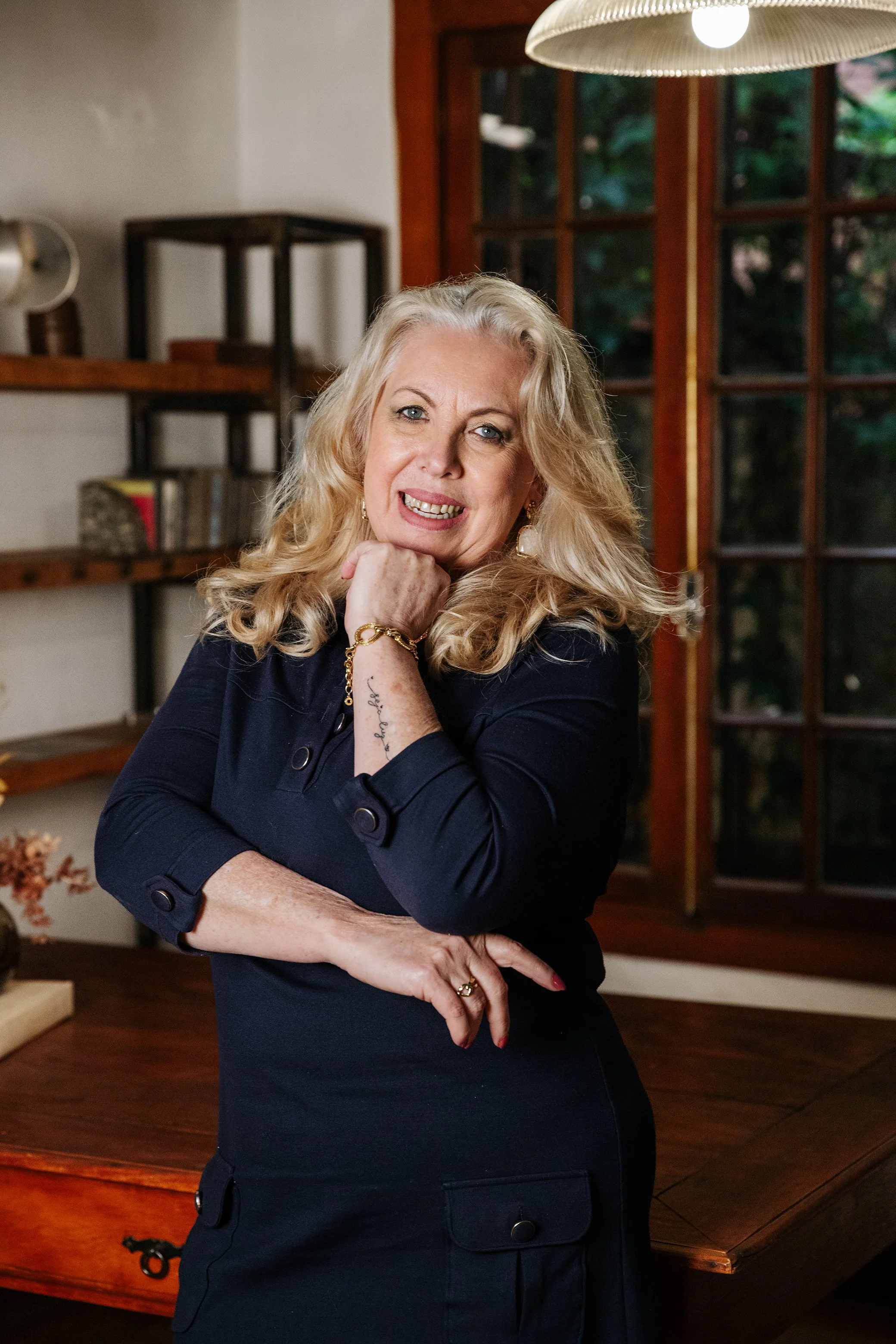 Mulher de cabelos loiros em um ambiente interno, usando roupa escura, com expressão facial confiante e gestos com as mãos, em um espaço decorado com prateleiras de madeira, livros e uma grande janela.