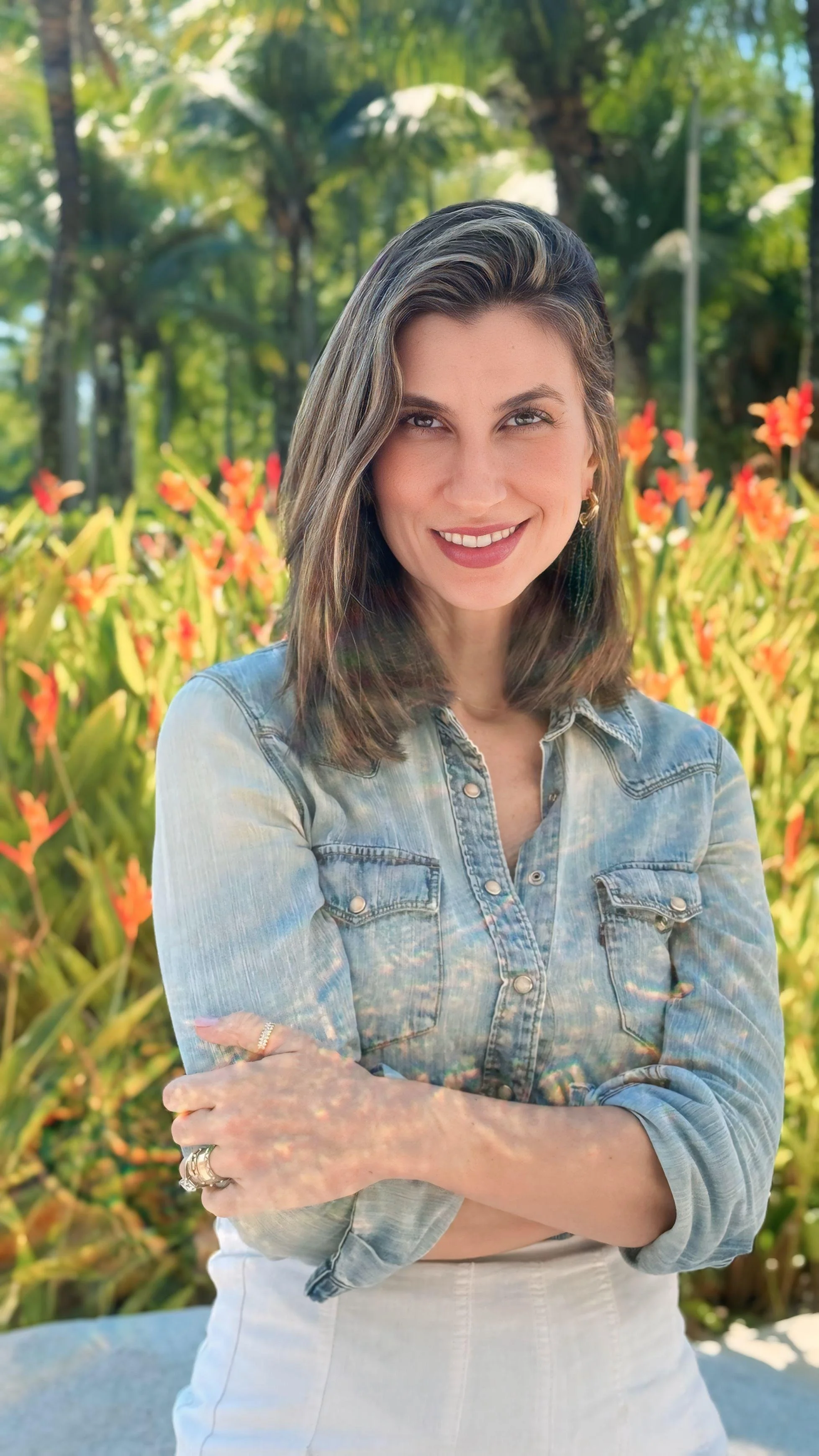 Mulher sorridente com cabelo castanho, usando jaqueta jeans clara e saia branca, posando ao ar livre com vegetação e flores ao fundo.
