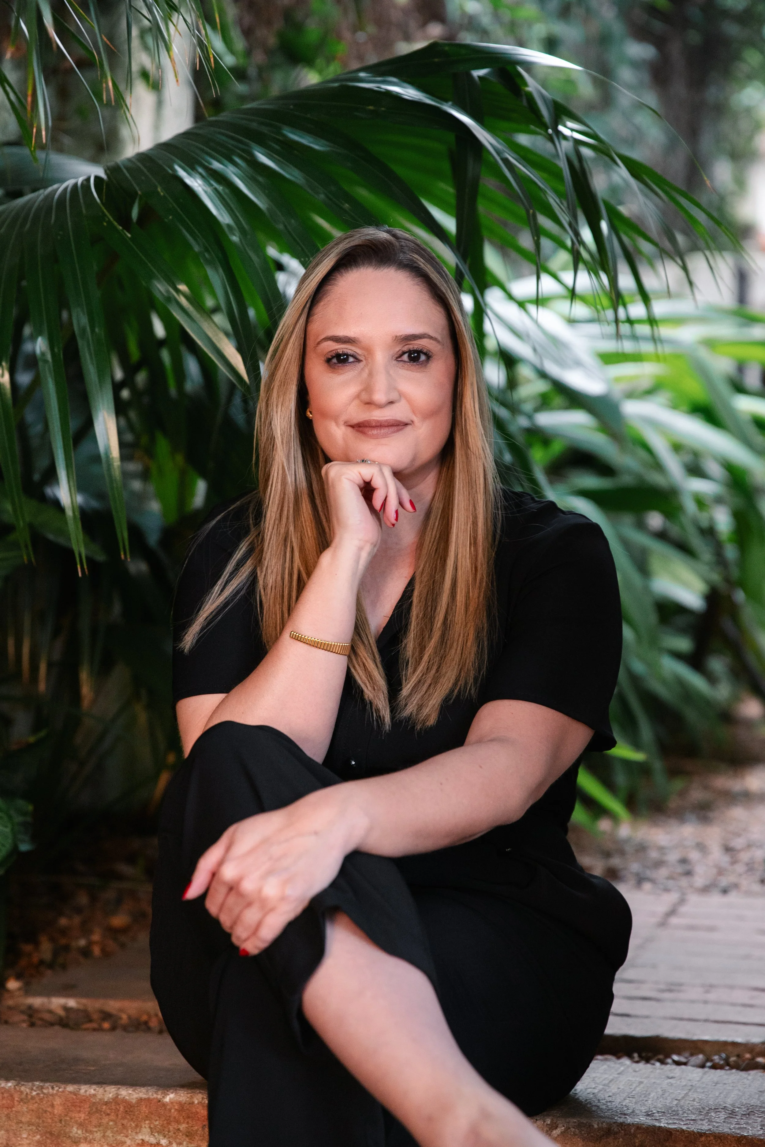 Mulher com cabelo castanho claro, vestindo roupa preta, sentada em uma superfície de pedra, cercada de plantas tropicais verdes, com expressão serena e olhando para a câmera.