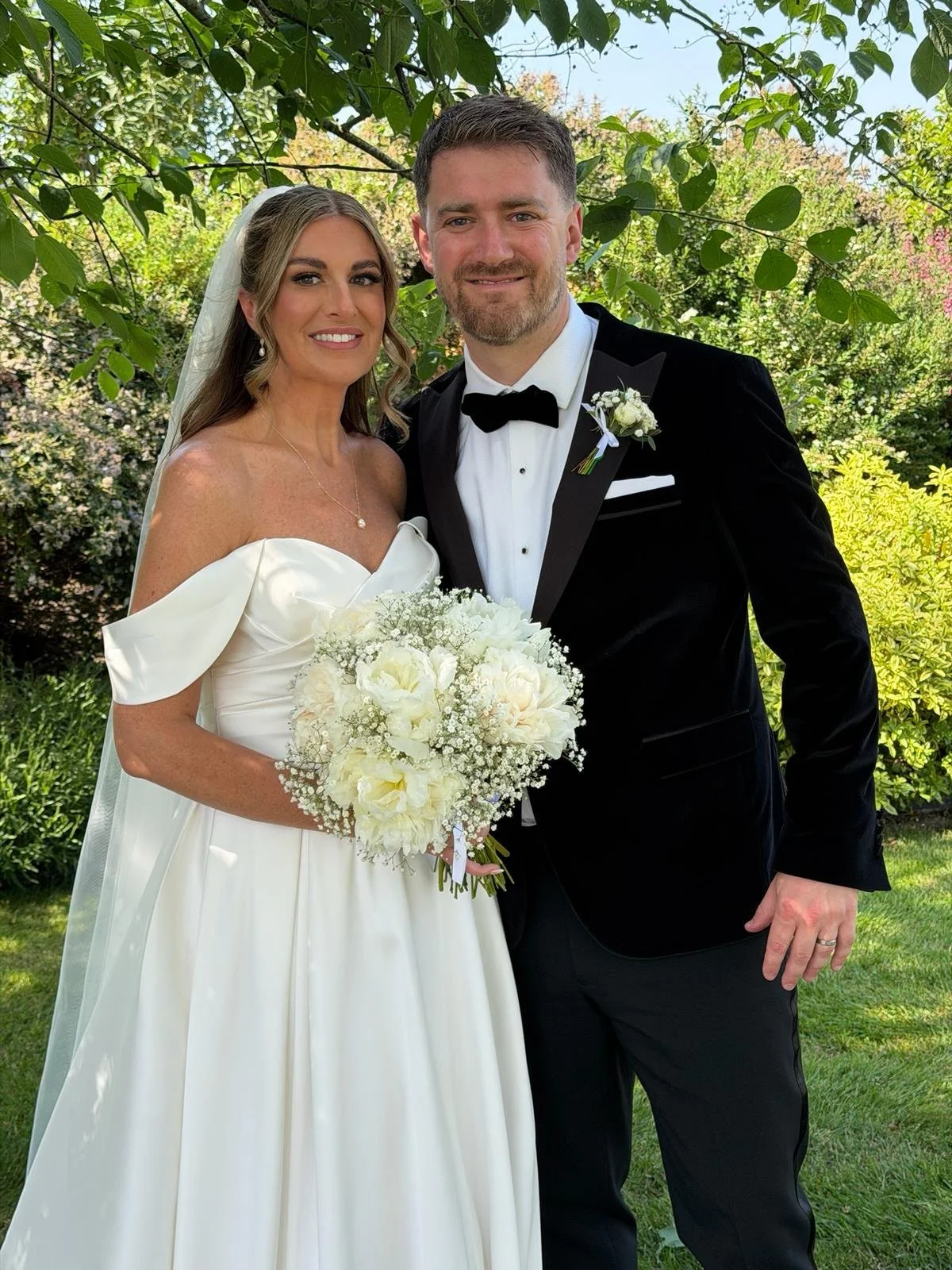 A bride and groom standing outdoors on their wedding day, smiling. The bride is holding a bouquet of white flowers and wearing a white off-the-shoulder wedding dress with a veil. The groom is dressed in a black tuxedo with a black bow tie, white shirt, and a boutonniere.