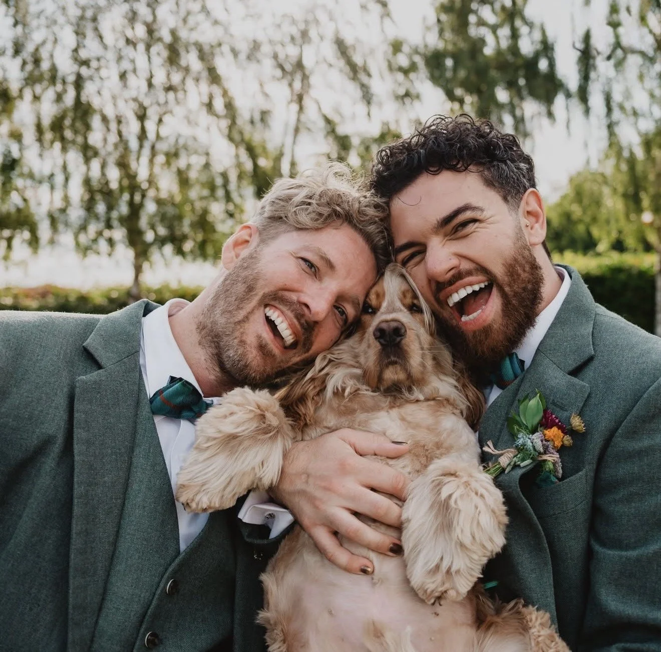 Two men in suits and bow ties holding a dog, smiling and leaning in together outdoors with trees in the background.