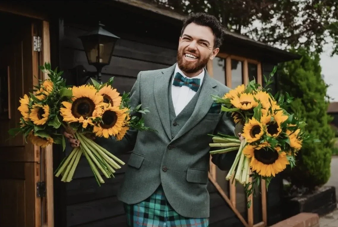 A man in a gray suit with a blue bow tie, holding two large bouquets of sunflowers, standing outside near a rustic wooden building with green trees nearby.