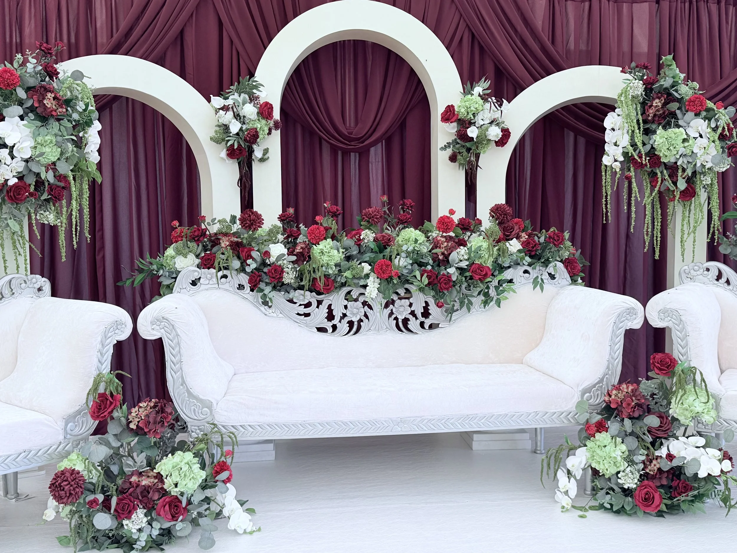 Floral wedding backdrop with white arches, burgundy and white flowers, and dark red curtains.