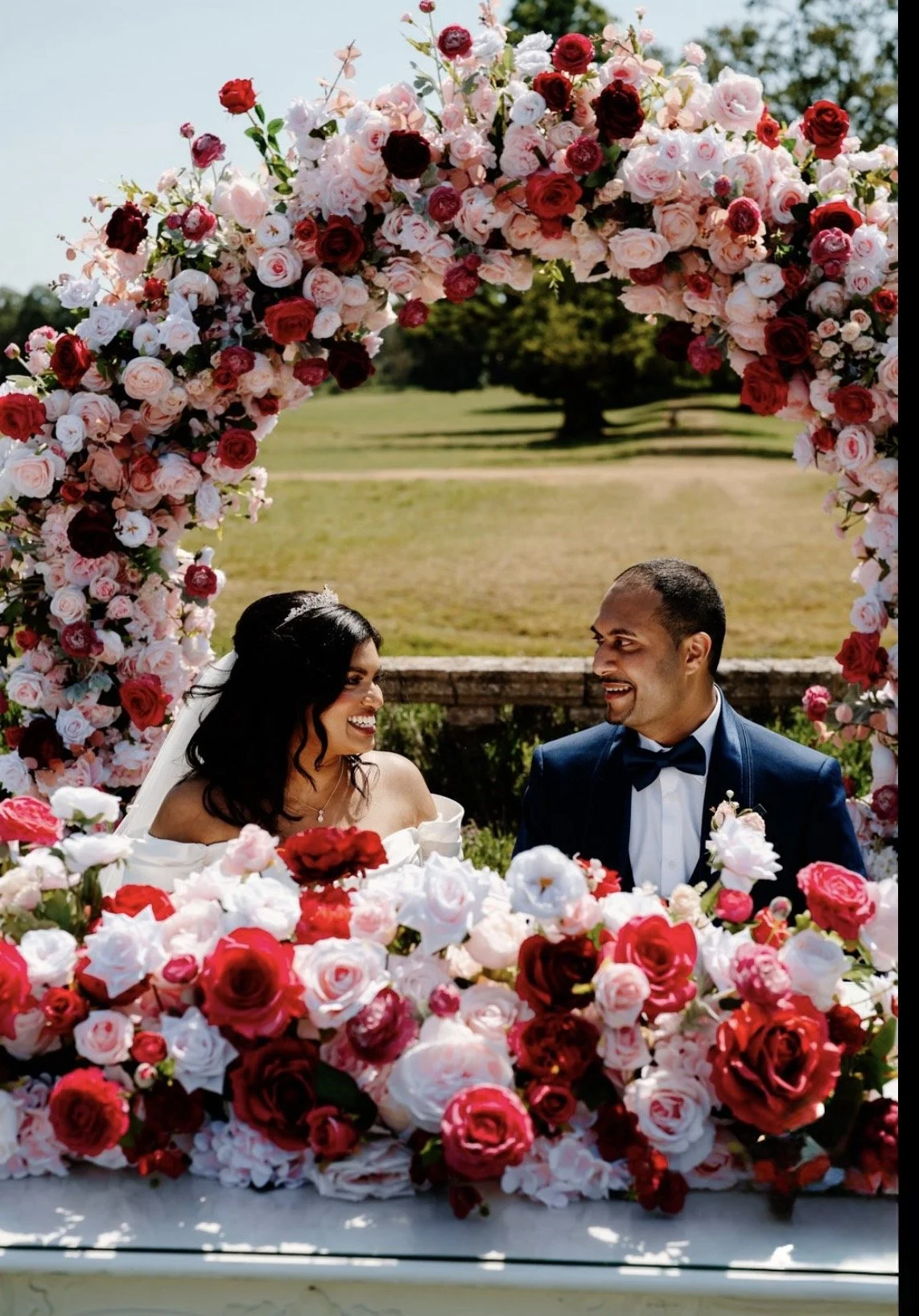 A bride and groom smiling at each other under a floral arch with pink and red roses on a sunny day outdoors.
