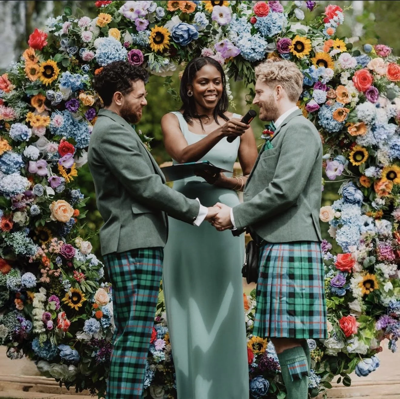 Two men in kilts holding hands and exchanging vows during a wedding ceremony, with a female officiant holding a microphone and a clipboard, standing in front of a large floral arch outdoor setting.
