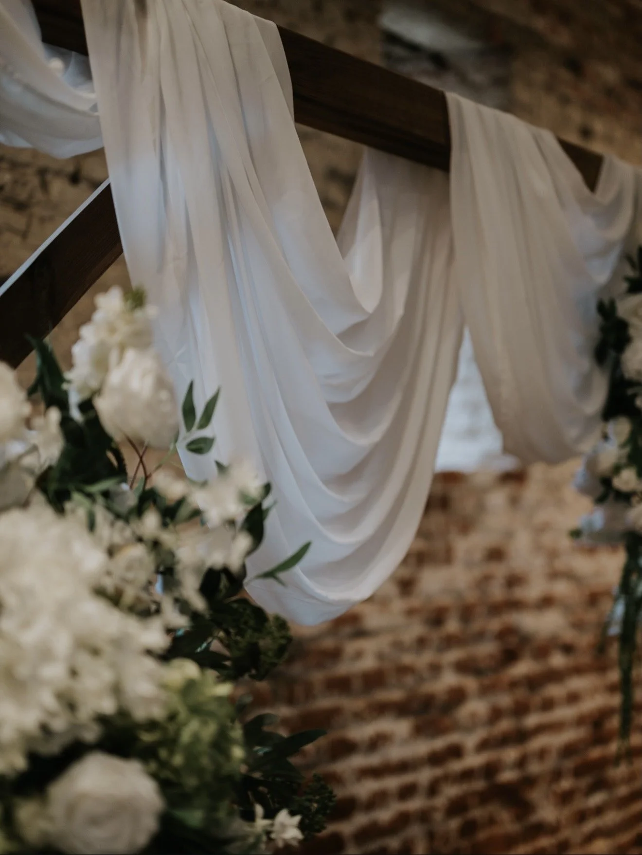 White drapes or fabric hanging from a wooden beam, decorated with green leaves and white flowers, possibly part of a wedding set-up.