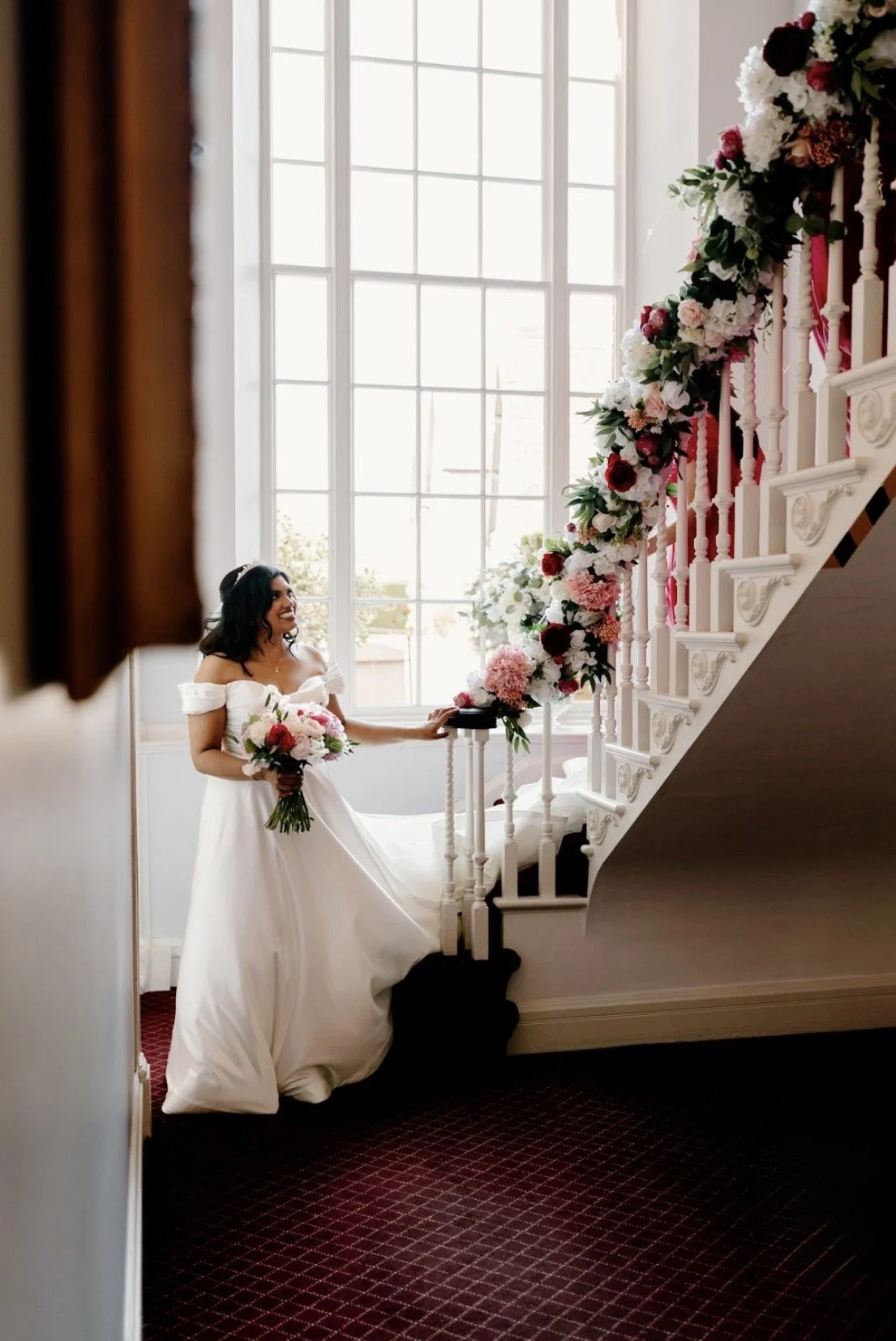 A bride in a white wedding gown holding a bouquet, standing near a staircase decorated with red and white flowers, inside a bright room with large windows.