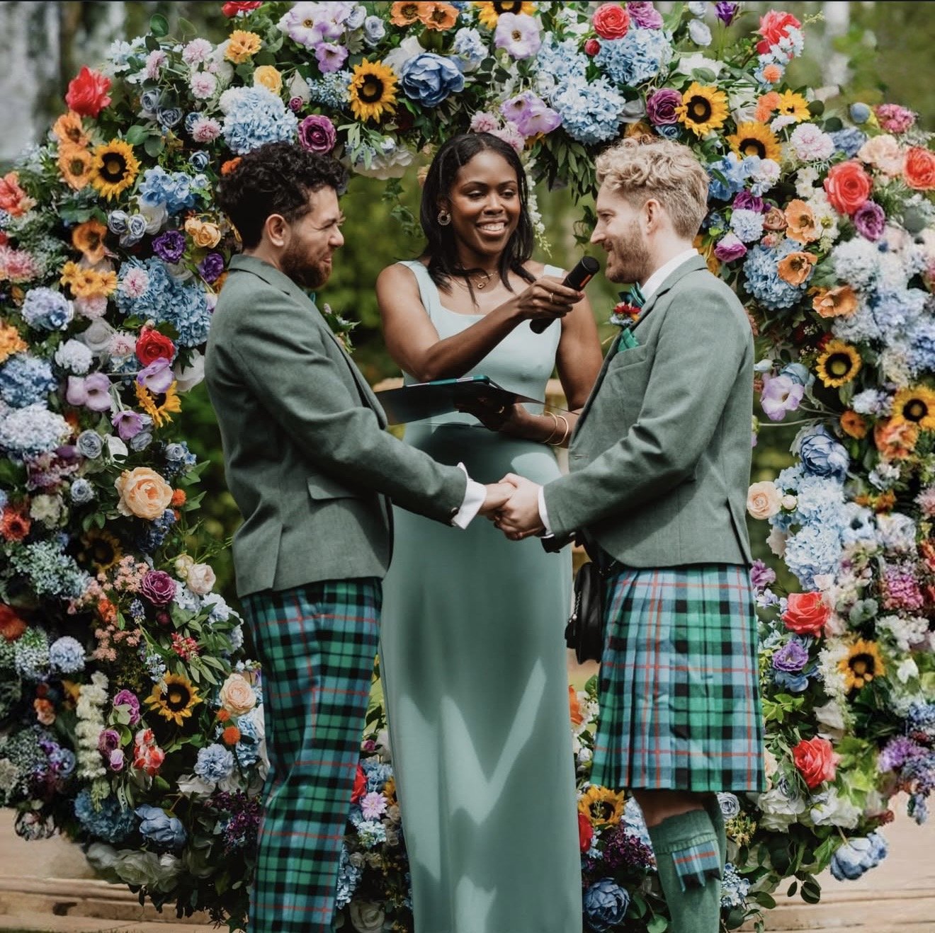 Two men in matching green tartan kilts, gray jackets, and white shirts hold hands during a wedding ceremony. A woman in a light blue dress officiates, holding a microphone and a clipboard. They stand under a floral arch made of colorful flowers.