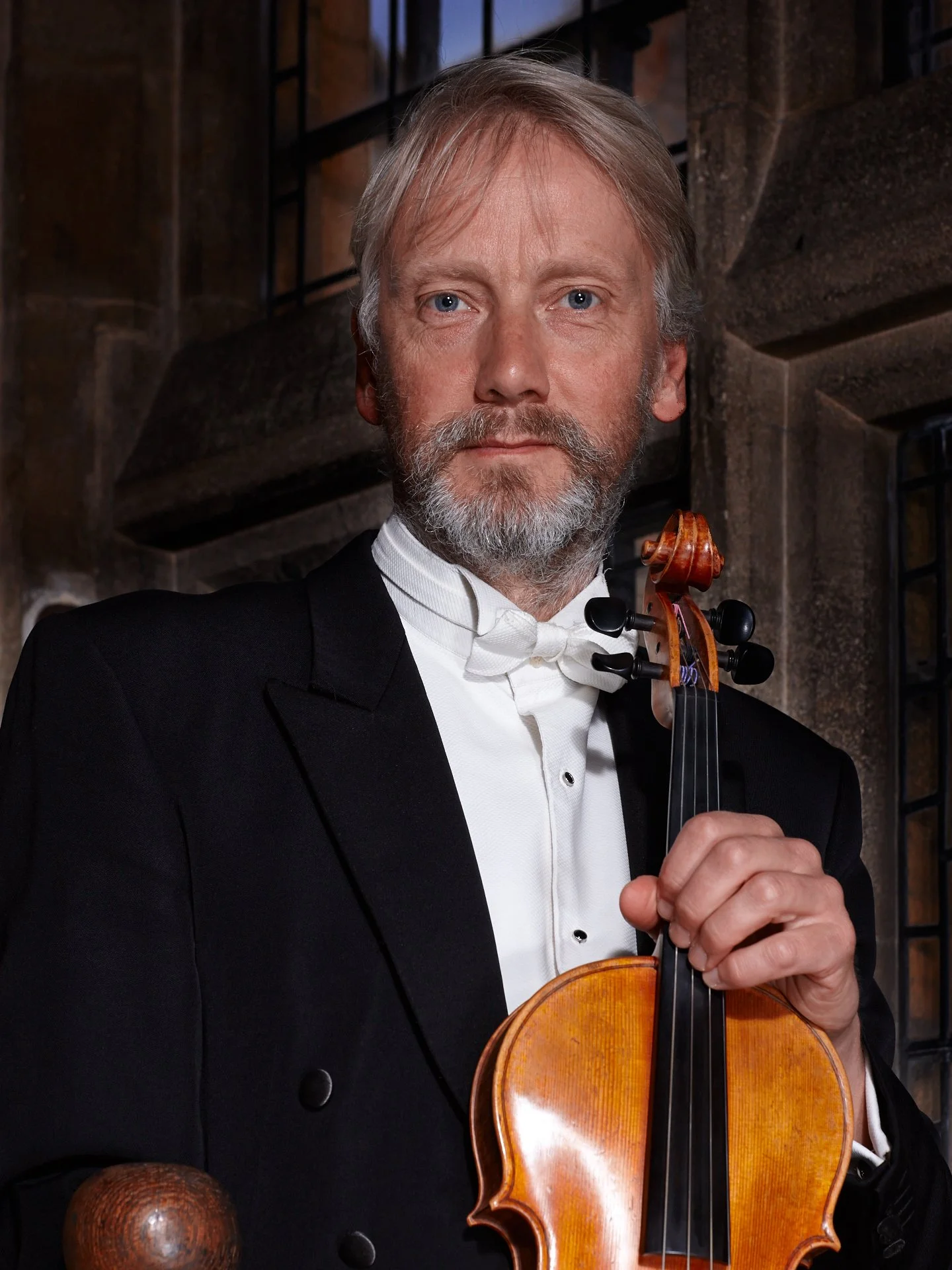 A man in formal attire holding a violin and bow, standing in front of a historical stone building with stained glass windows.