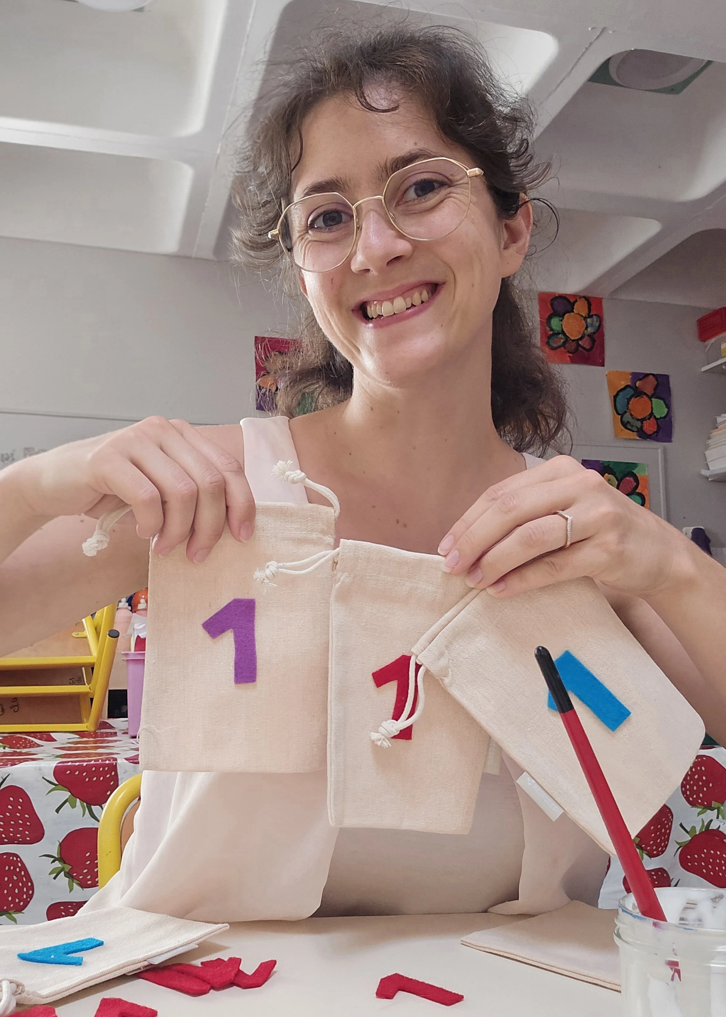Une femme souriante portant des lunettes, tenant des pochons avec des chiffres colorés (chiffre 1). Elle se trouve dans une classe d'école: une salle décorée de peintures colorées d'animaux. Elle est fière de montrer ses pochons colorés.