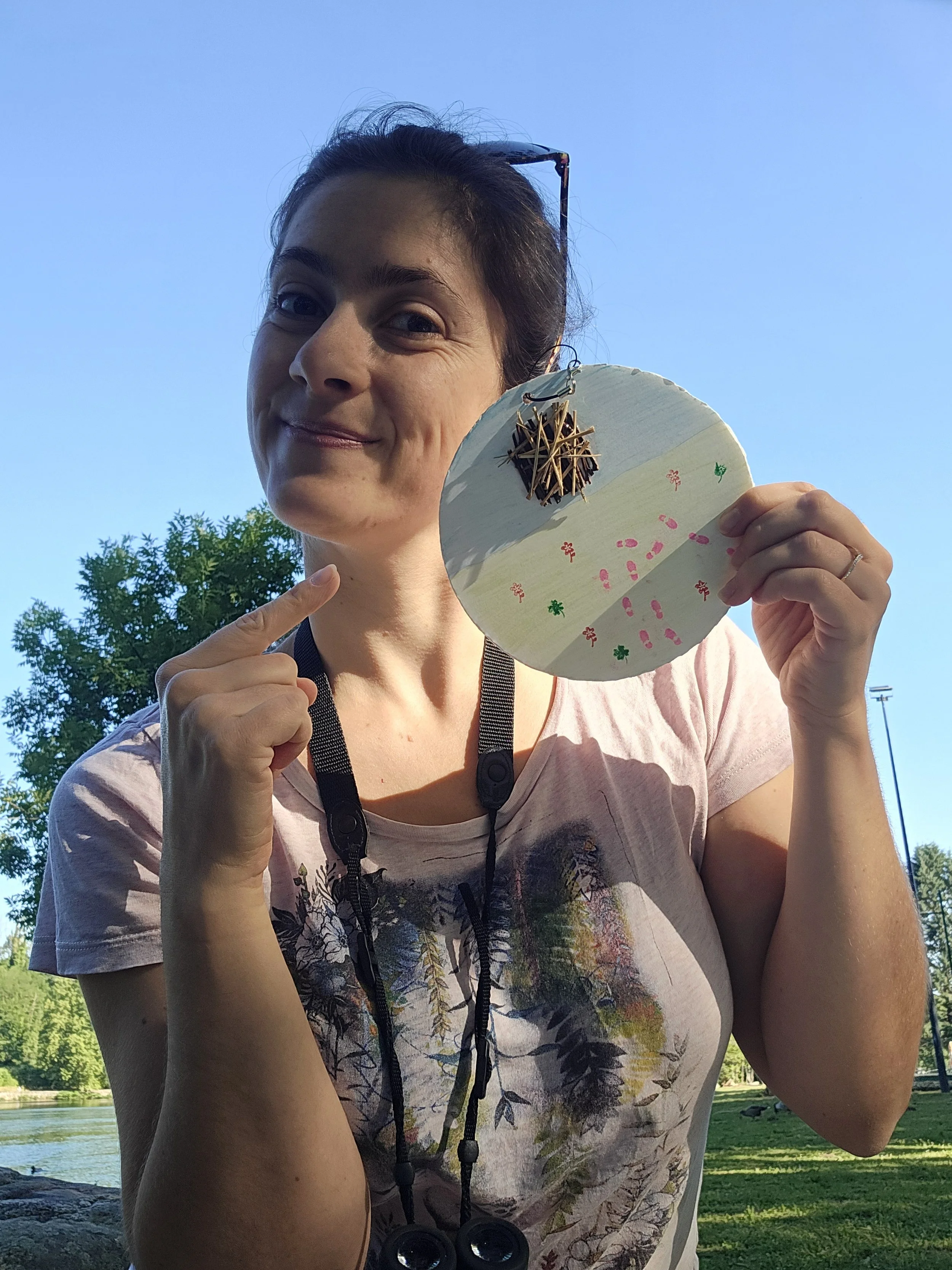 Femme souriante tenant un cercle en carton décoré de petits motifs illustrant un lac et un nid d'oiseau. Elle est en extérieur près d'un lac avec des arbres et un ciel clair. Elle porte des jumelle autour du cou et des lunettes de soleil sur la tête.