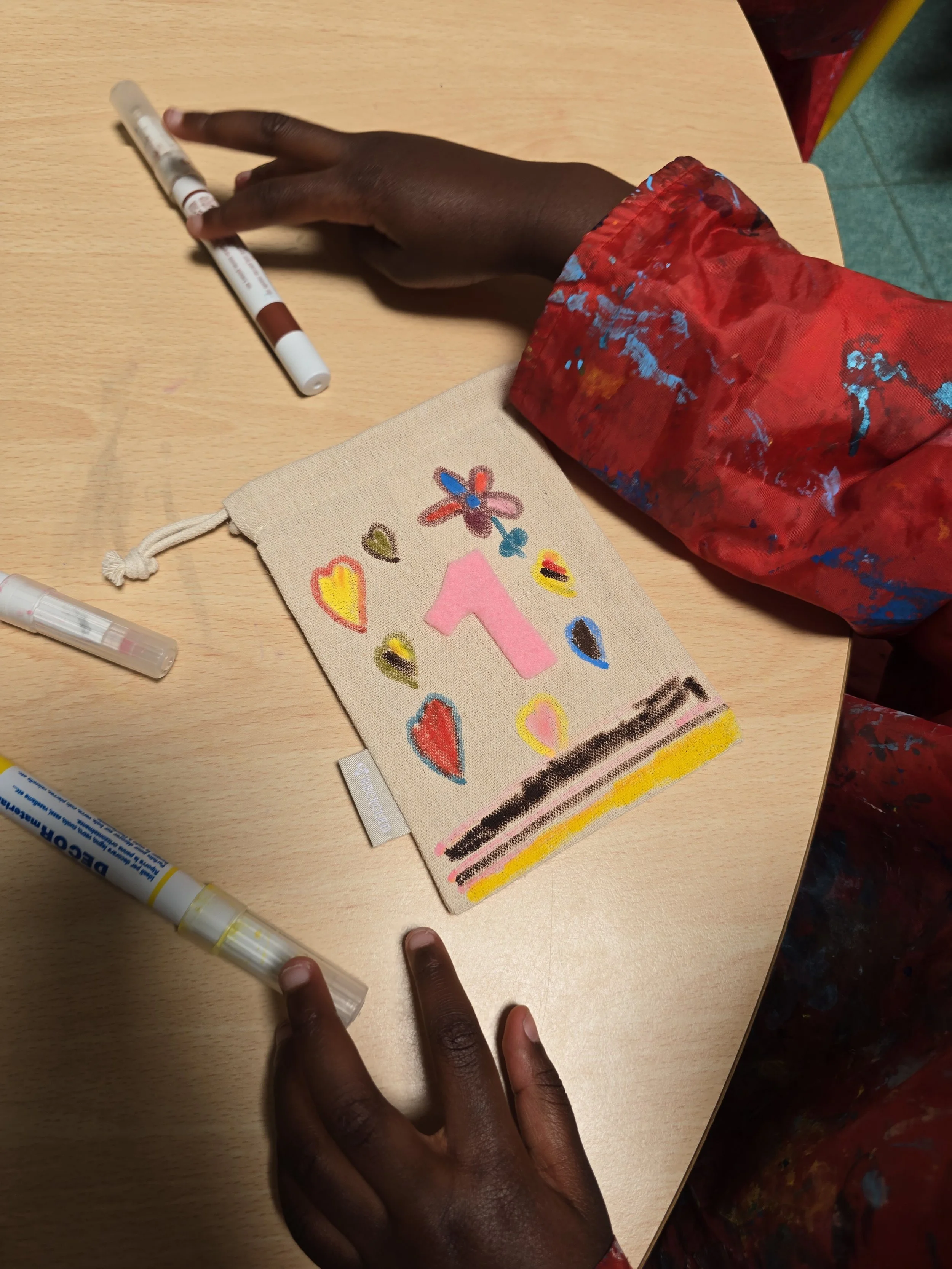 Une main noire portant une manche rouge avec des taches de peinture, posée sur une table en bois clair, avec un stylo à peinture et un sac en toile beige orné de dessins colorés de cœurs, fleurs et autres motifs. L'enfant décore le sac.