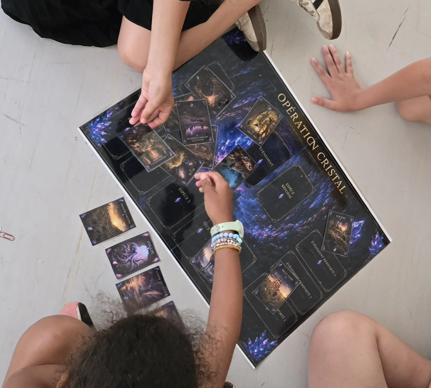 Groupe d'enfants jouant à un jeu de société appelé "Opération Cristal" au sol, avec plusieurs cartes posées devant eux et un plateau de jeu sombre.
