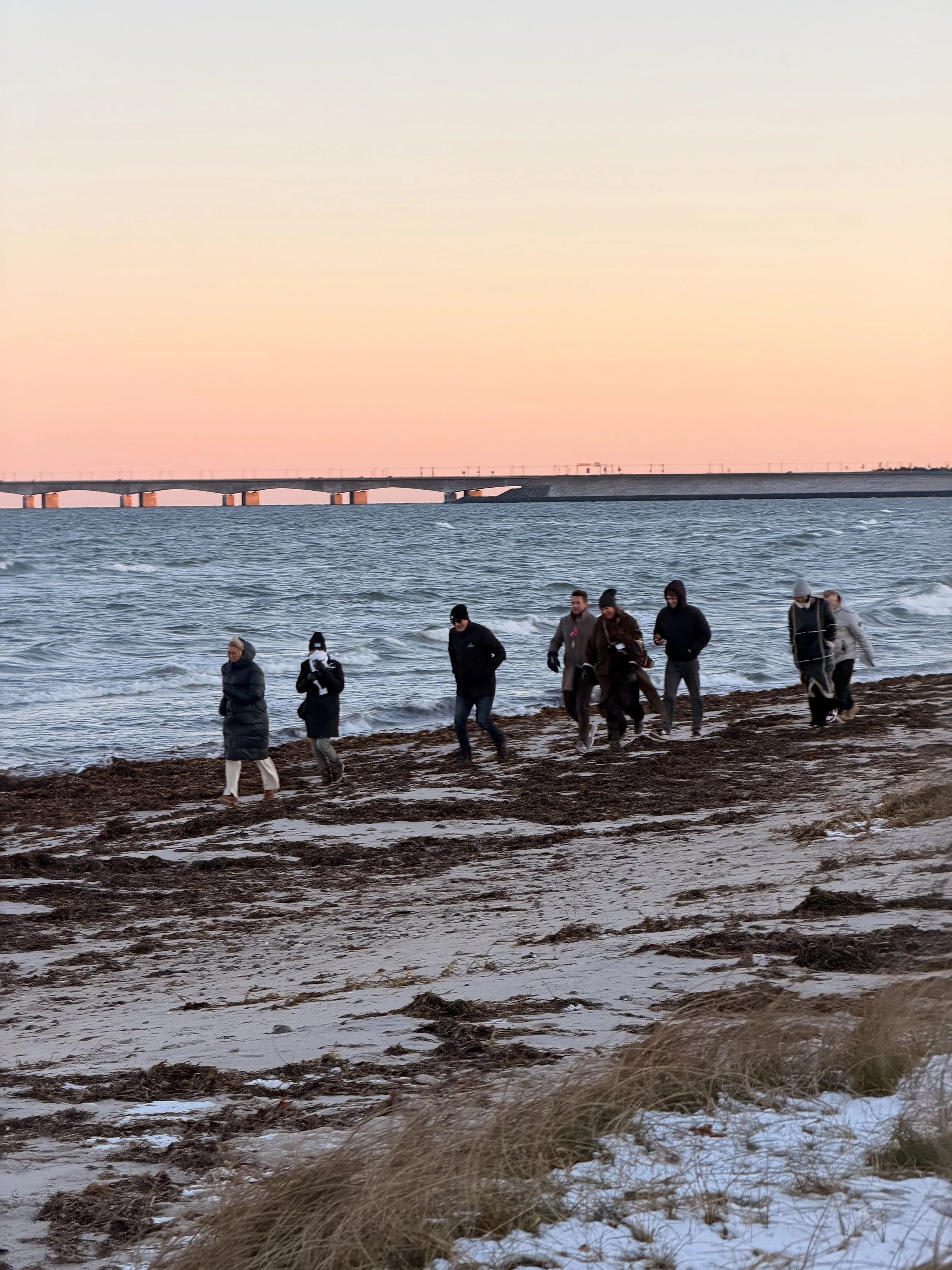 Gruppe mennesker går langs en strand med hav og en bro i baggrunden ved solnedgang.