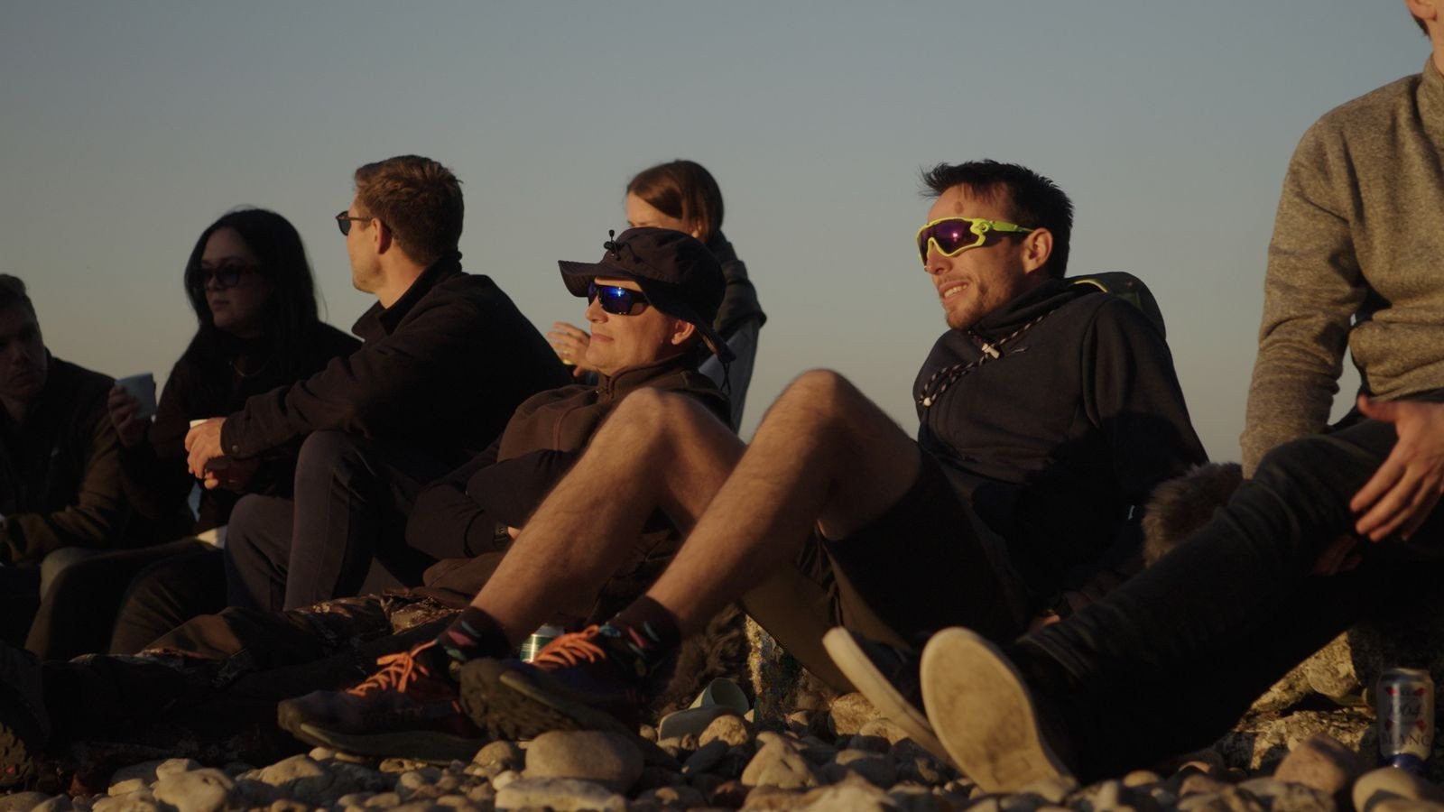 Gruppe venner sidder på en stenstrand, nogle med solbriller, nyder solnedgangen